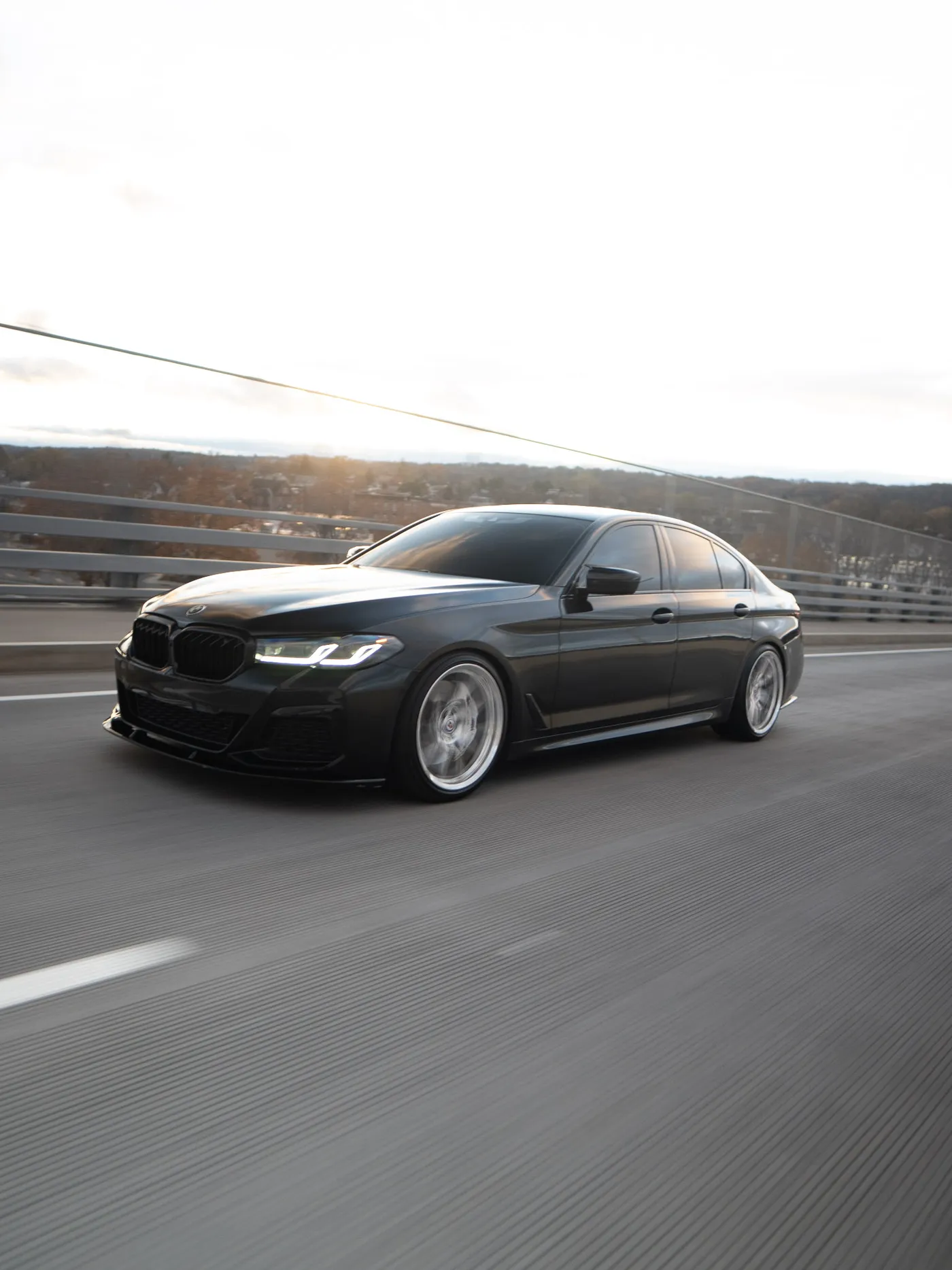 Black BMW sedan on a bridge in low sun, car slightly closer to camera with a lane line at lower left