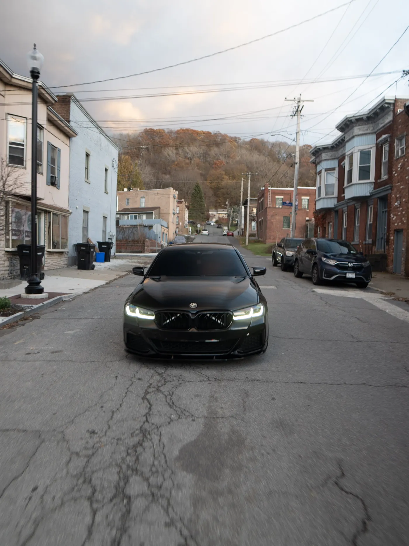 Black BMW sedan head-on on a cracked street with row houses and a wooded autumn hillside behind