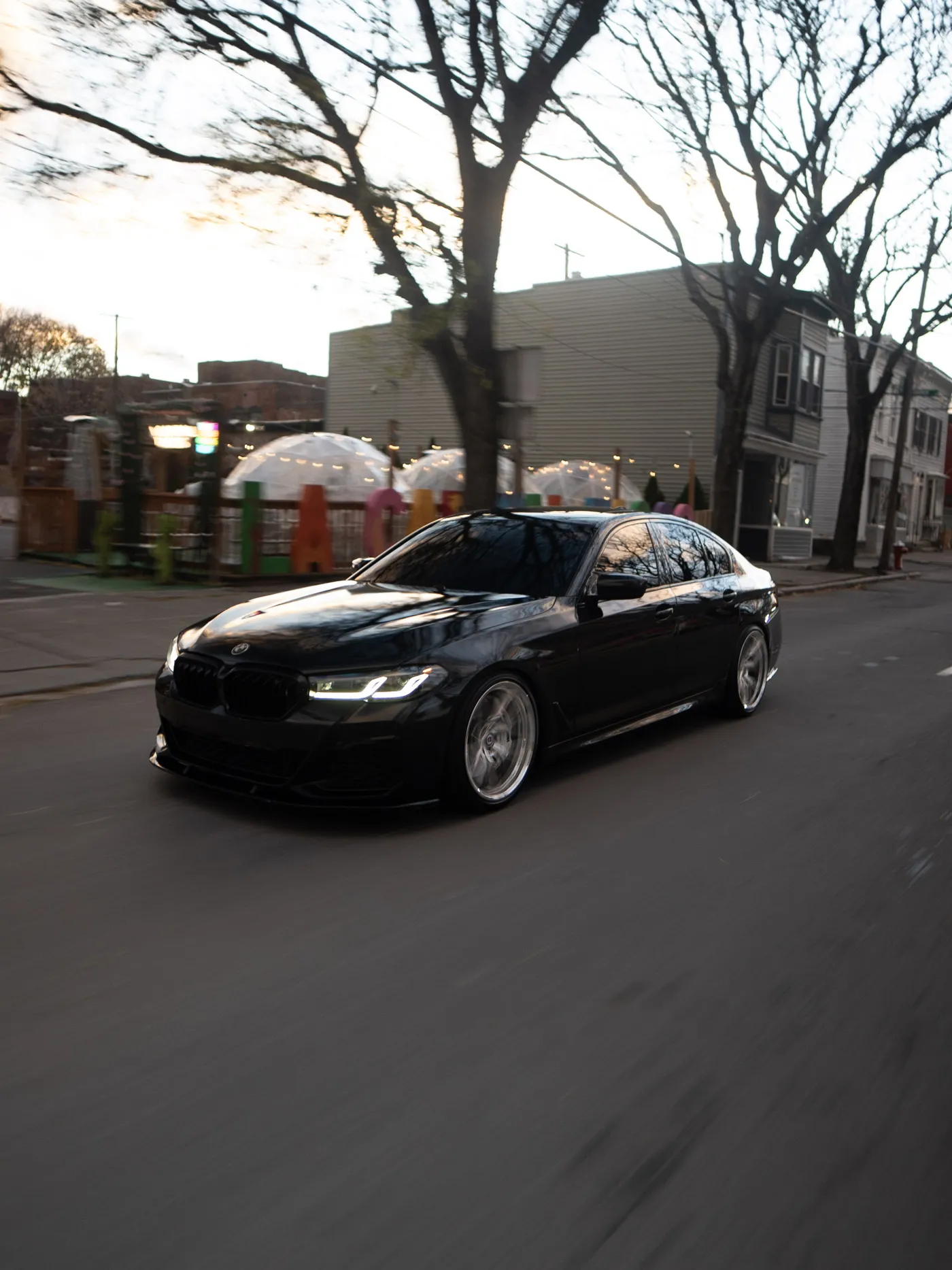 Black BMW sedan with silver wheels passing a fenced patio with white igloo domes and bare trees
