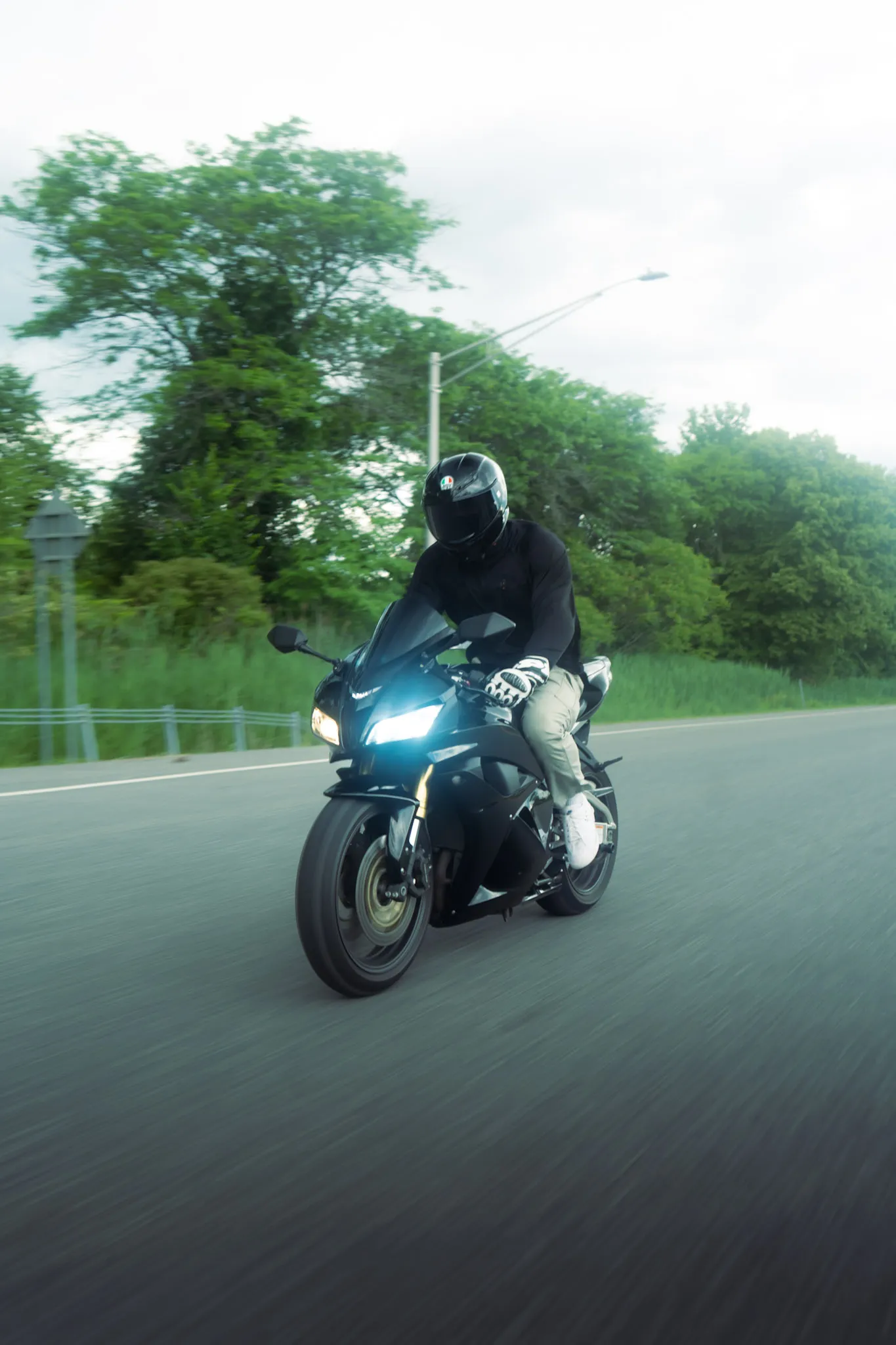 Rider on a black sport motorcycle with headlights on, riding toward the camera on a tree-lined road