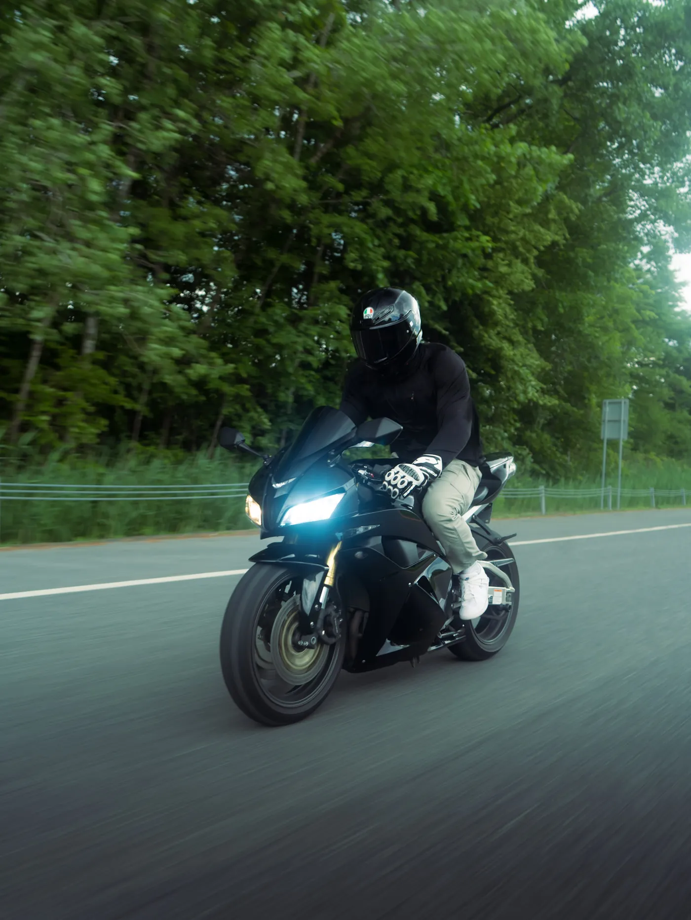 Rider in a black helmet on a black sport motorcycle with headlight on, riding along a tree-lined road