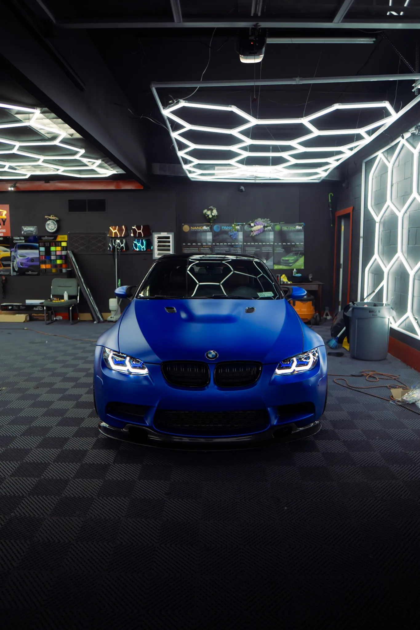 Matte blue BMW coupe seen head-on with headlights on, parked under hexagon ceiling lights in a tint shop bay