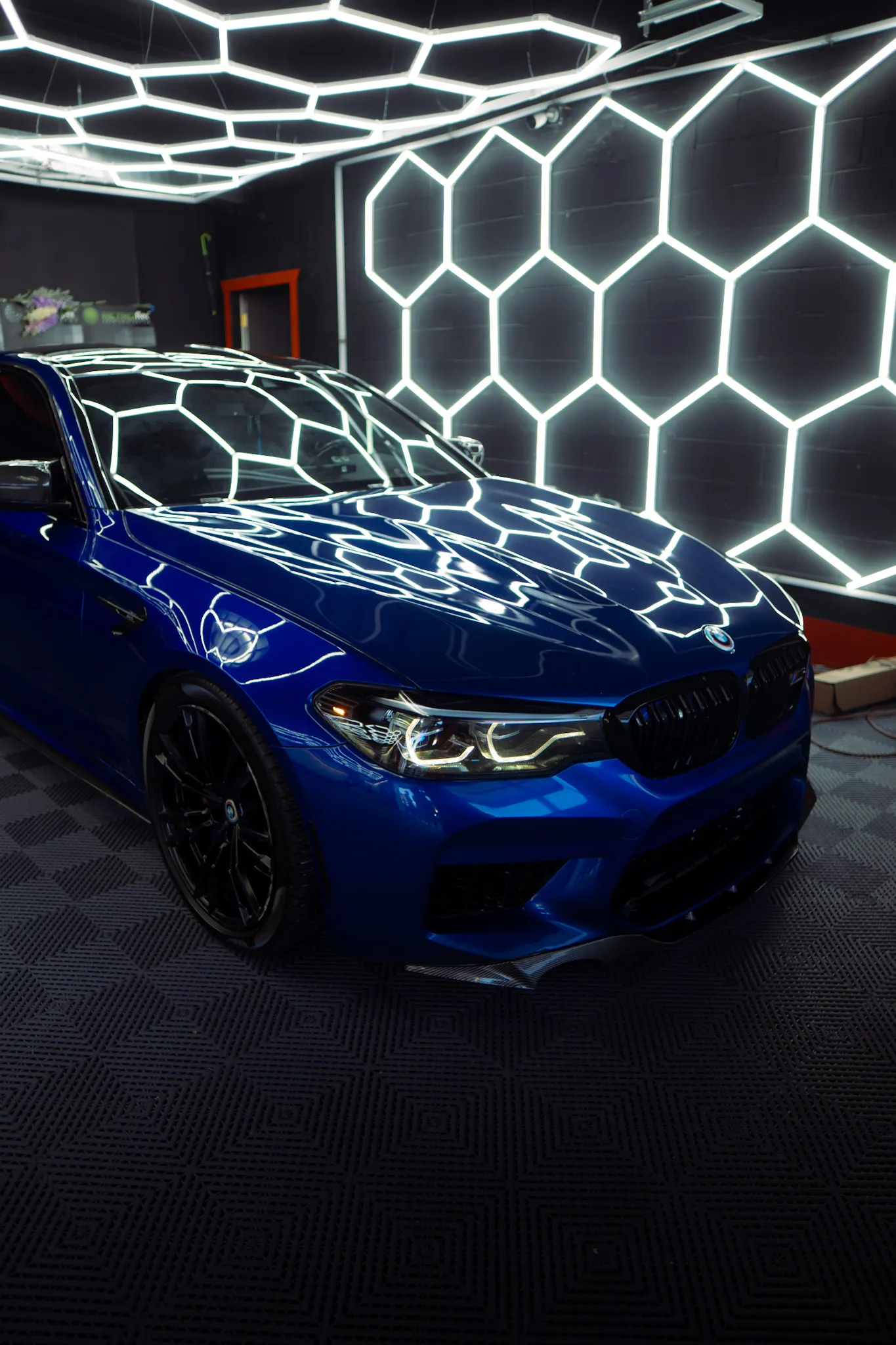 Front three-quarter view of a glossy blue BMW sedan with black wheels beside a hexagon light wall