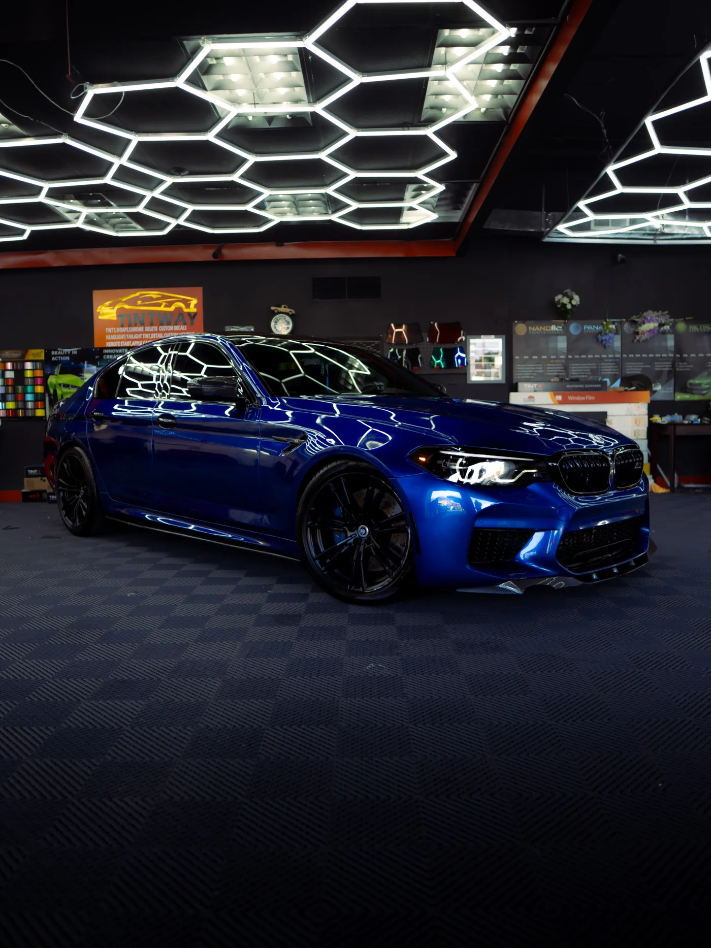Side view of a glossy blue BMW sedan in a tint shop with hexagon ceiling lights and film displays