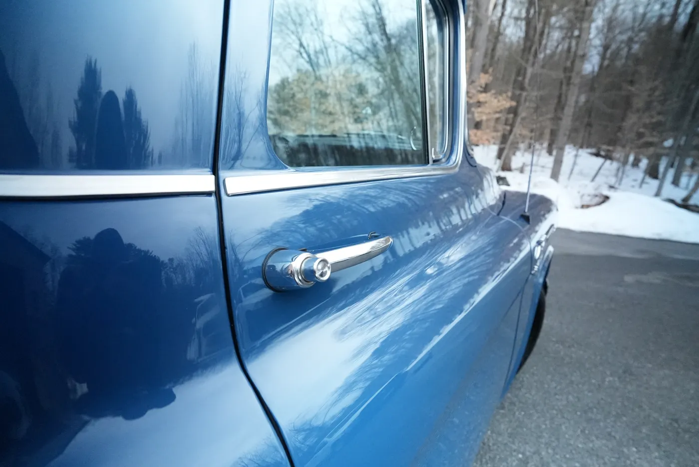 Close-up of the chrome door handle on a glossy blue classic pickup, snowy woods reflected in the paint