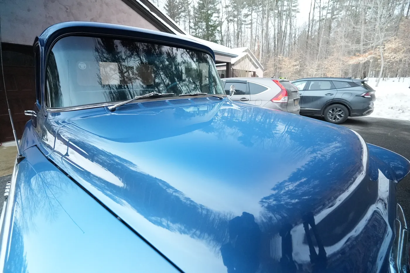 Close-up of the glossy blue hood of a classic truck reflecting trees, with a garage and two parked SUVs behind