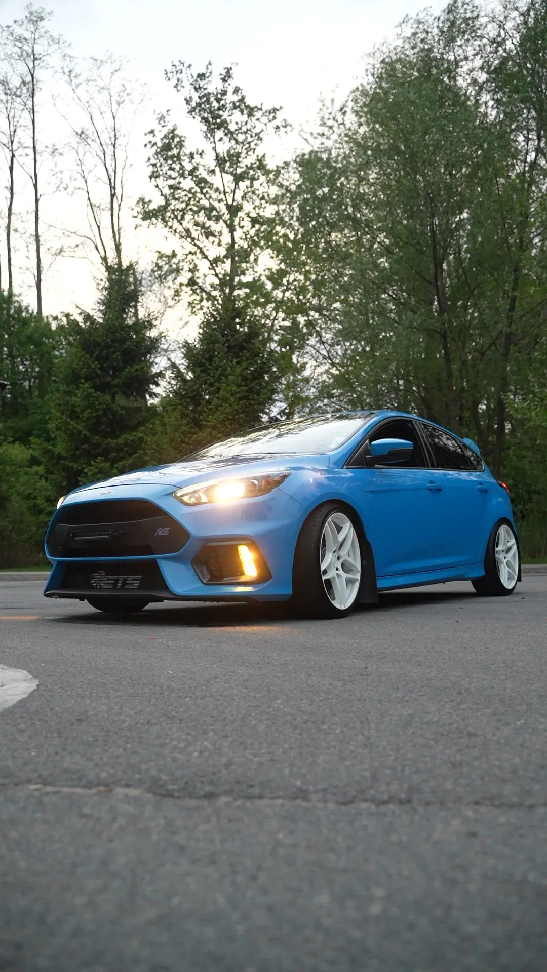 Blue Ford Focus RS with headlights on in a tree-lined parking lot