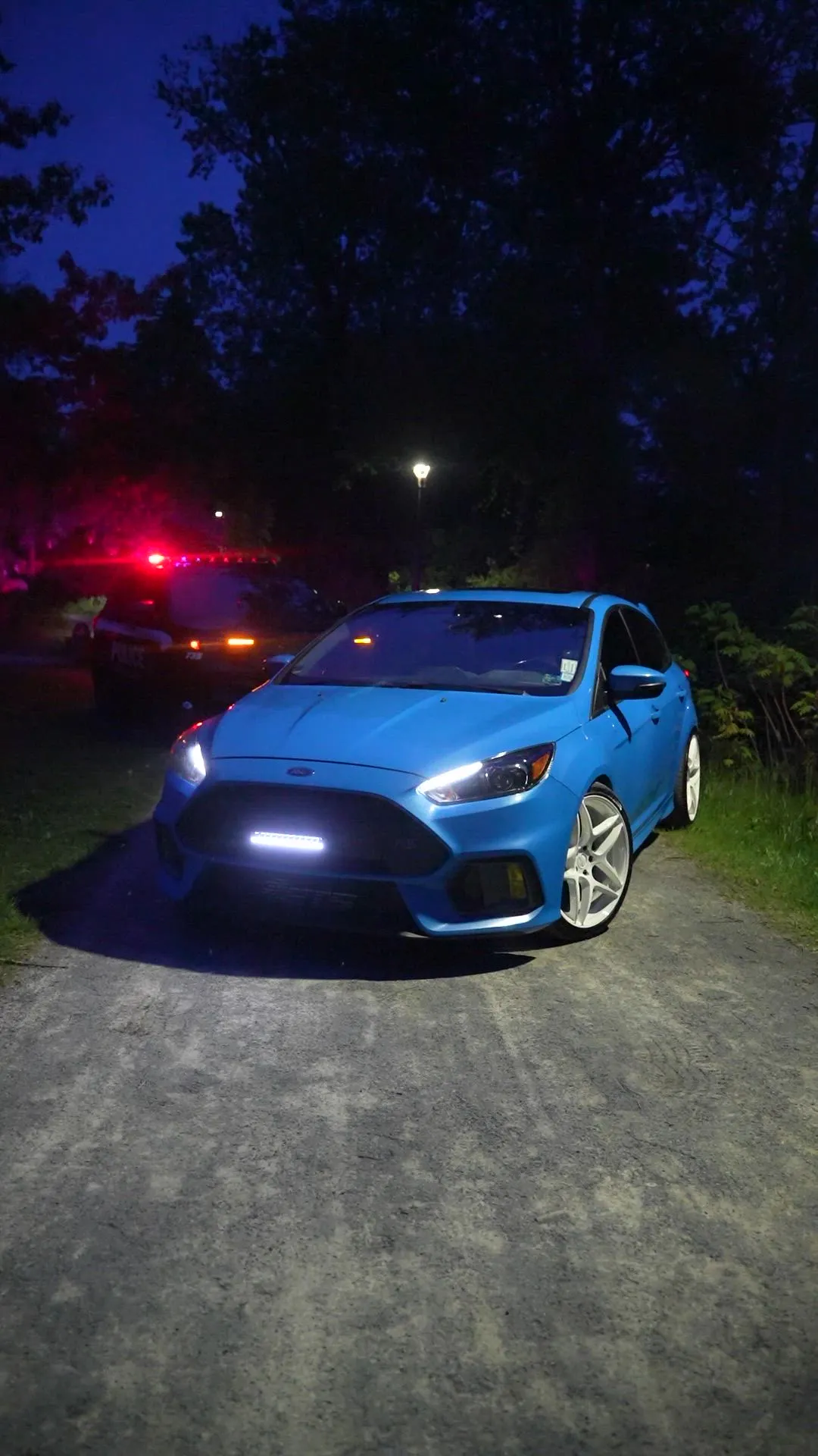 Front view of a blue Ford Focus RS with light bar on at night on a gravel path, red emergency lights behind