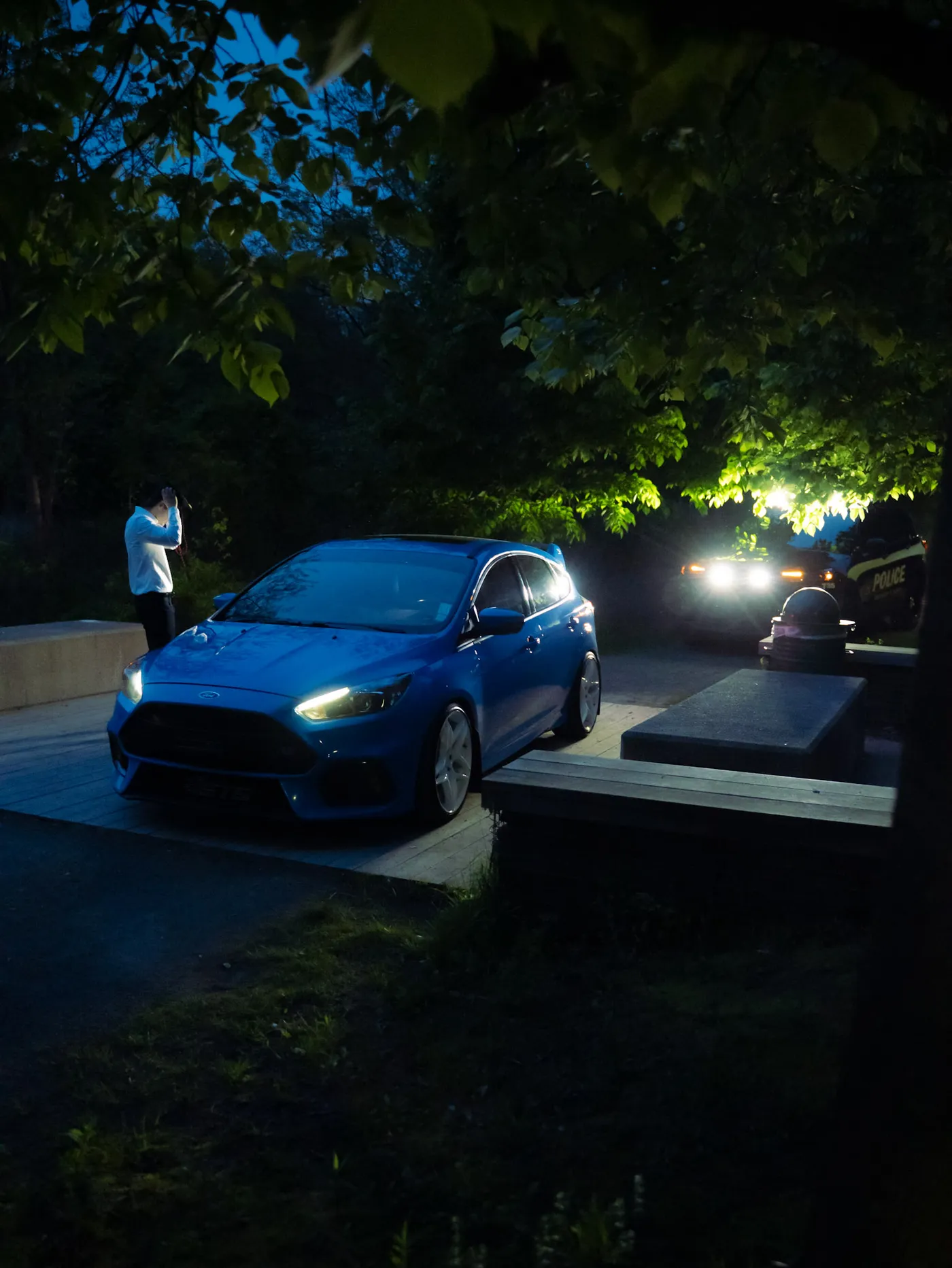 Blue Ford Focus RS parked under trees at night while a man photographs it, police vehicle lit in the background