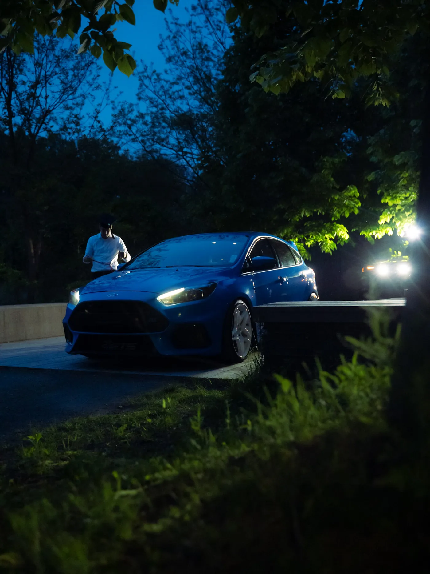 Blue Ford Focus RS with headlights on at night under trees, man in a white shirt looking down beside it