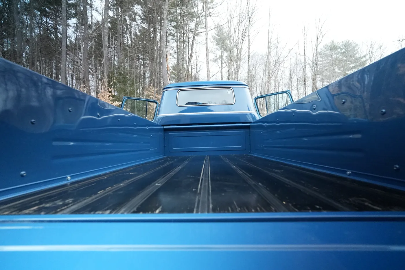 View along the empty bed of a blue classic pickup toward the cab with both doors open, bare trees behind