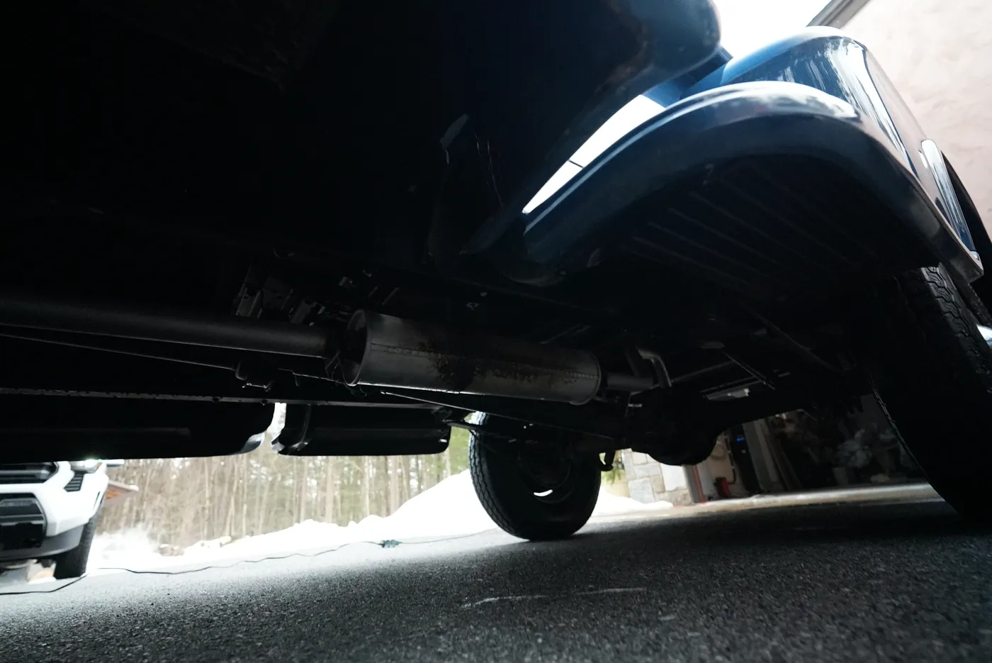 Ground-level view under a blue classic pickup showing the exhaust, rear axle and tyre on a driveway, snow behind