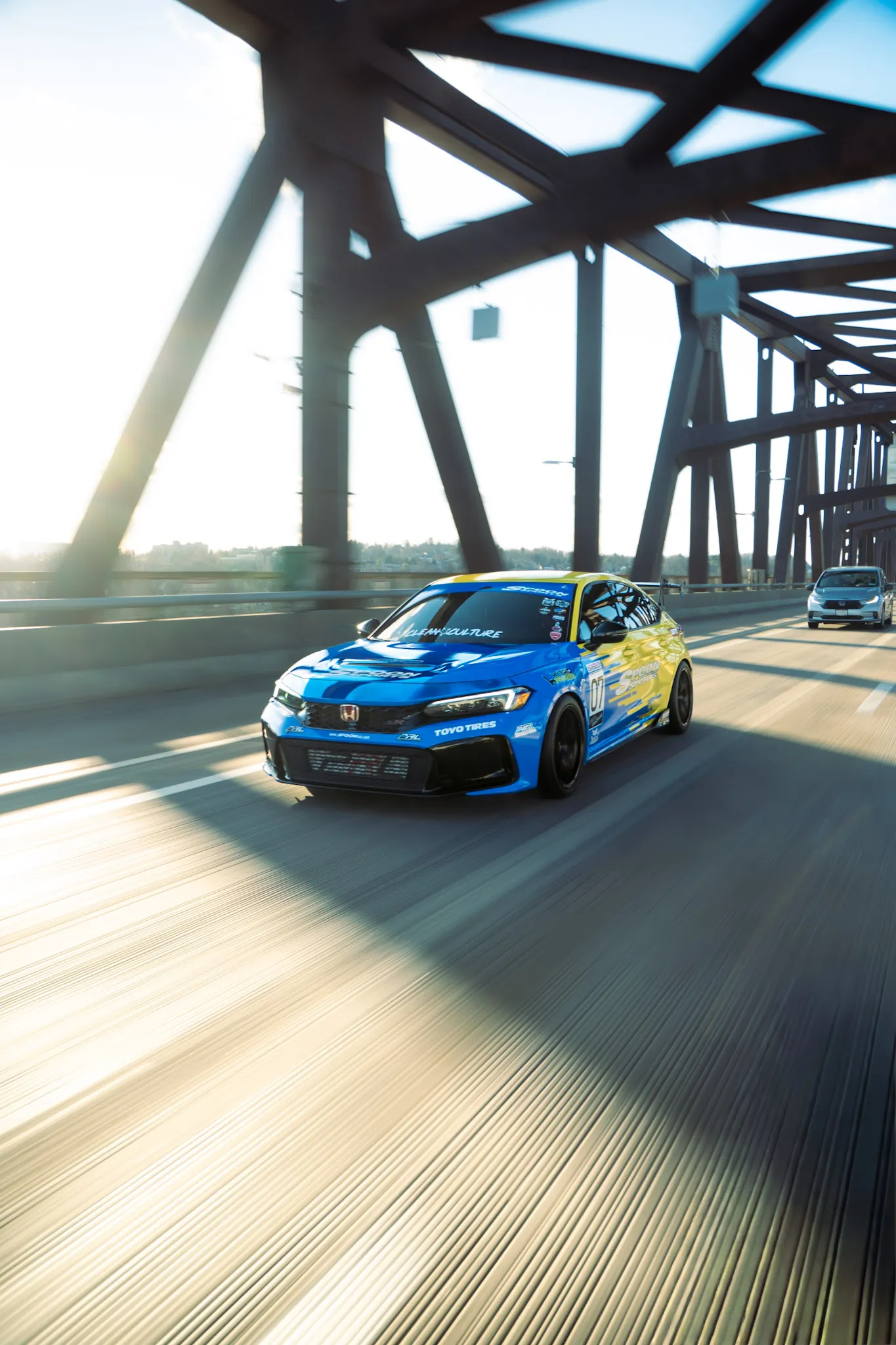 Blue and yellow Honda Civic driving centred under a steel truss bridge, silver minivan following behind