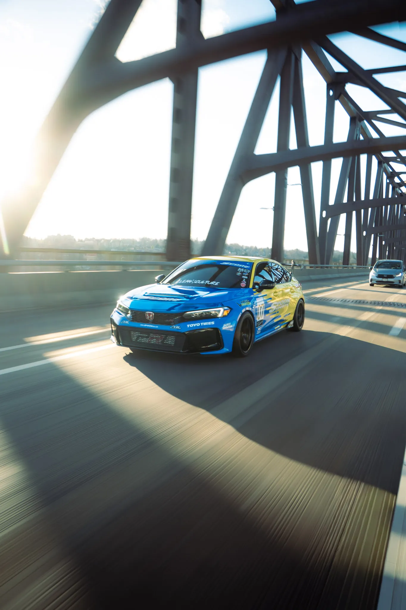 Blue and yellow Honda Civic crossing a steel truss bridge with bright sun flare on the left