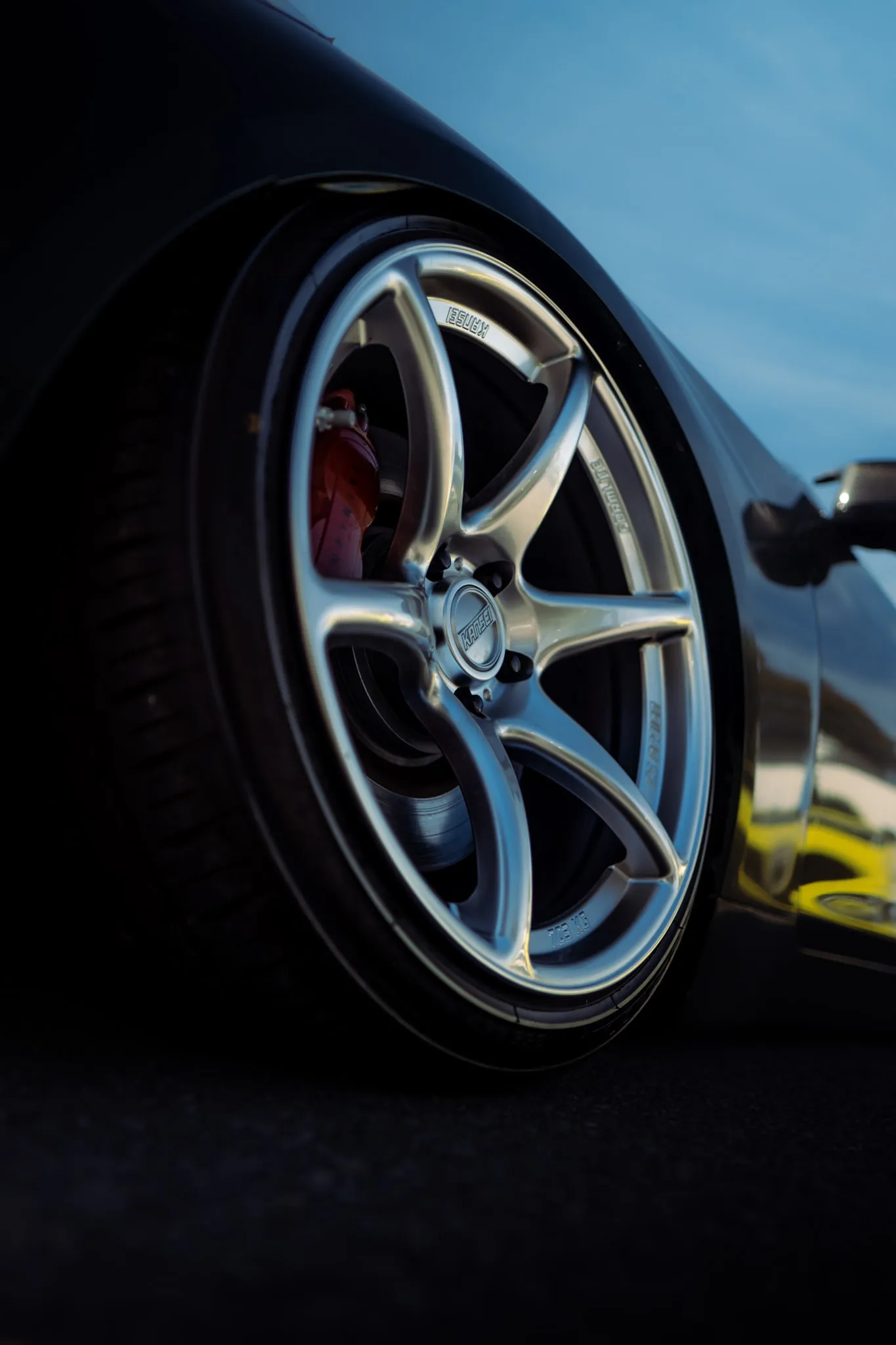 Silver six-spoke wheel with red caliper on a dark lowered car, shot from ground level outdoors under blue sky