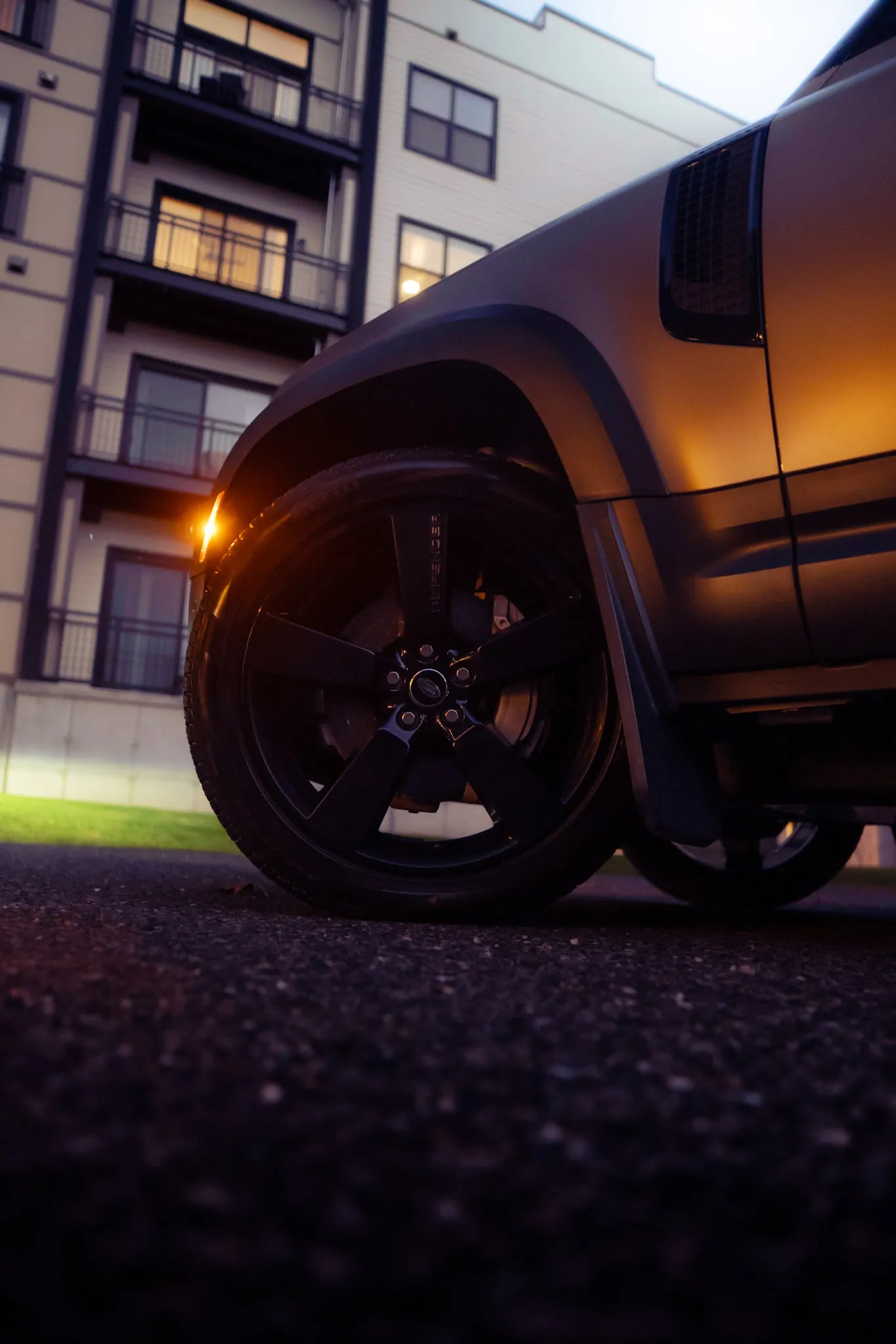 Ground-level close-up of a Land Rover Defender front wheel and fender vent, apartment balconies lit behind
