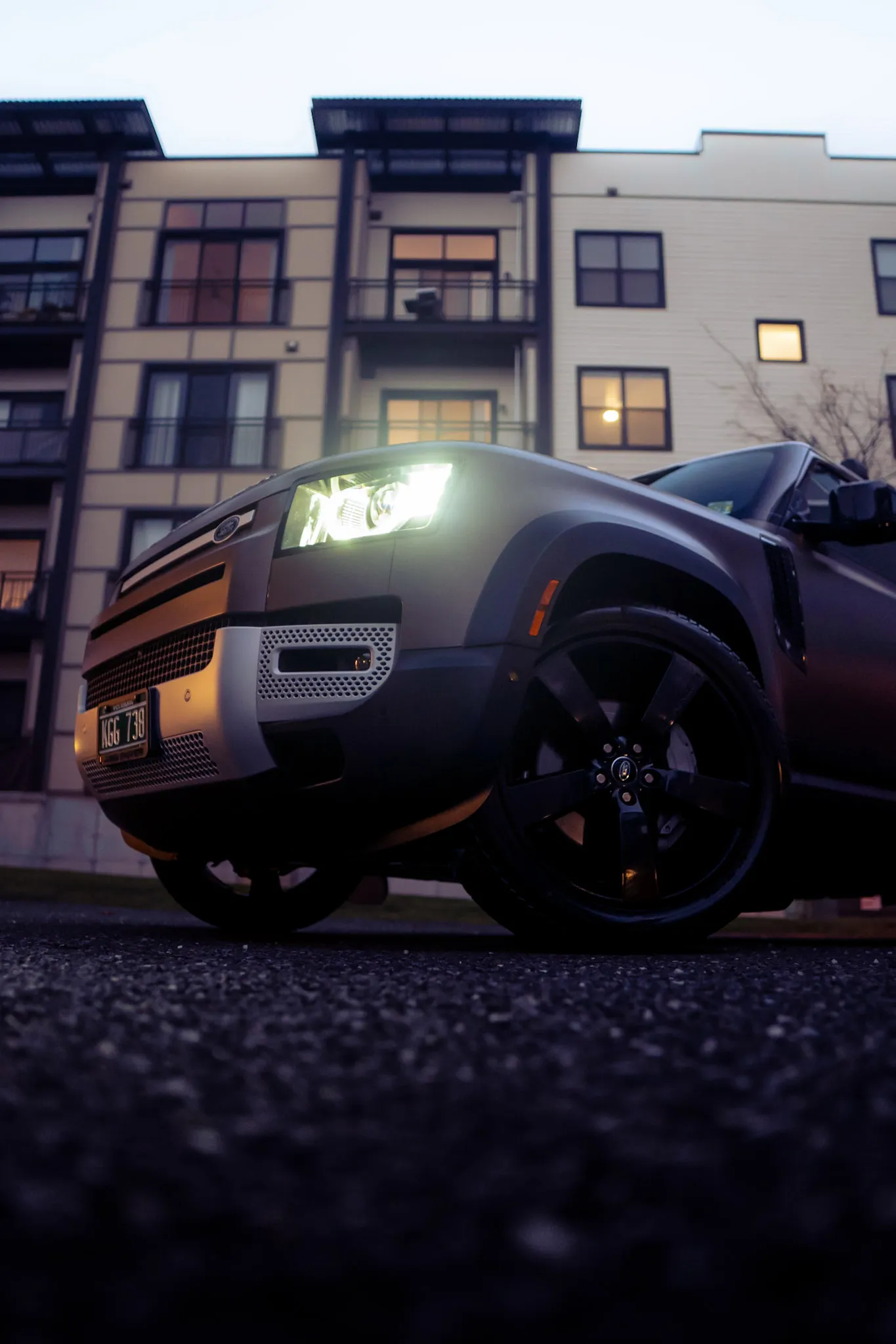 Ground-level close view of a matte grey Land Rover Defender's glowing headlight, front bumper and black wheel