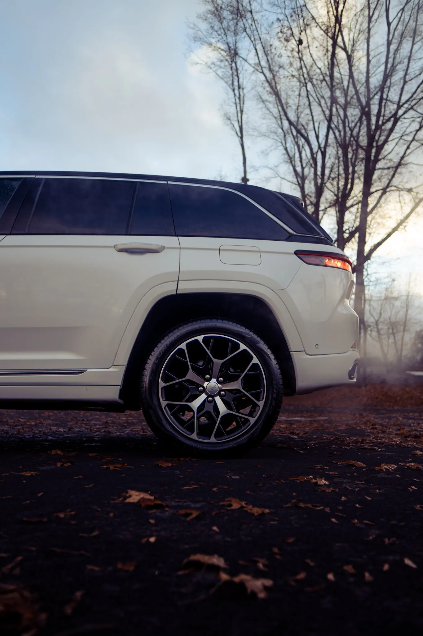 Side view of the rear wheel, rear door and lit tail light on a white Jeep Grand Cherokee, bare trees behind