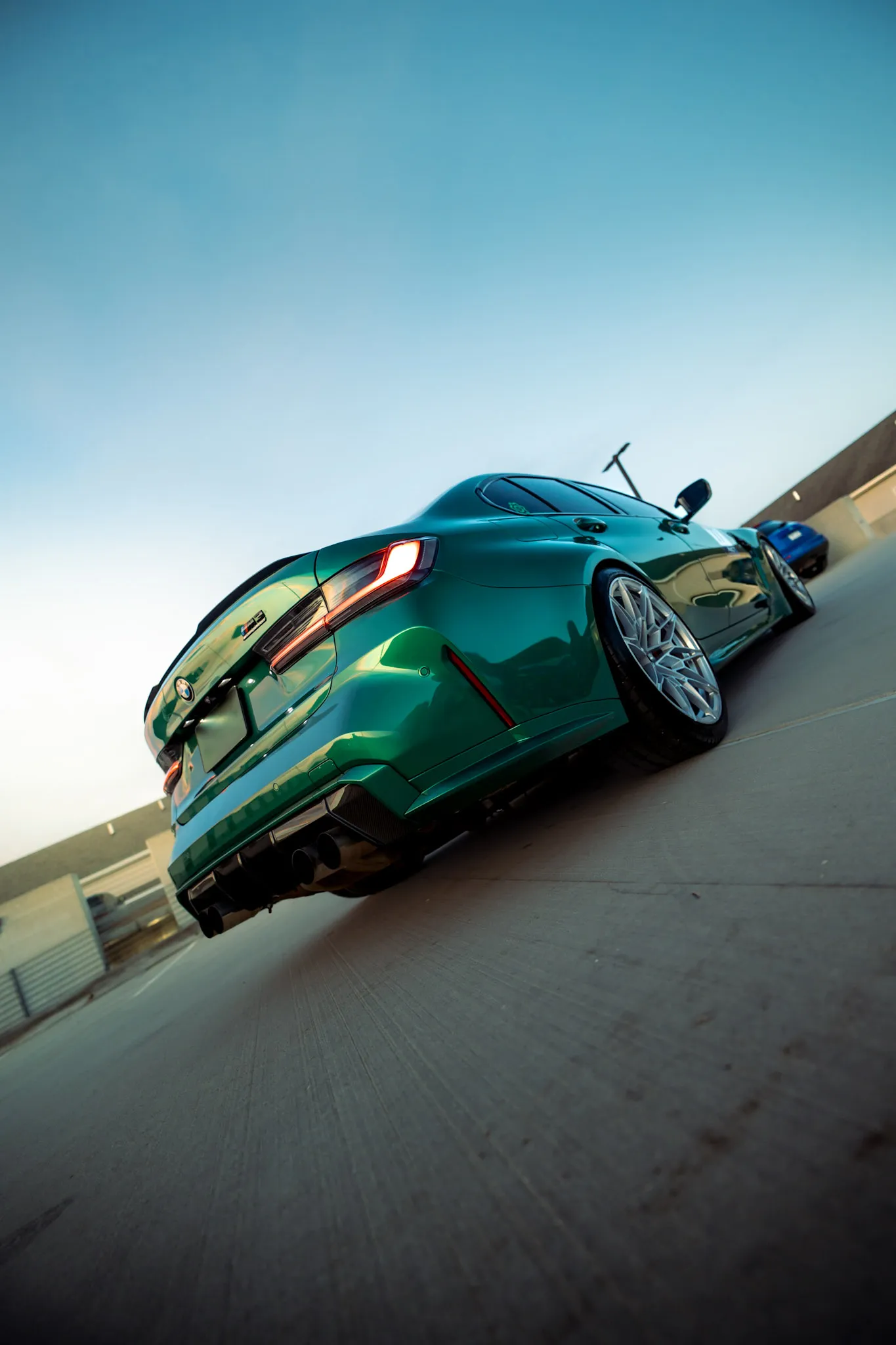 Low tilted rear three-quarter of a green BMW sedan with a blue car parked beyond, clear blue sky