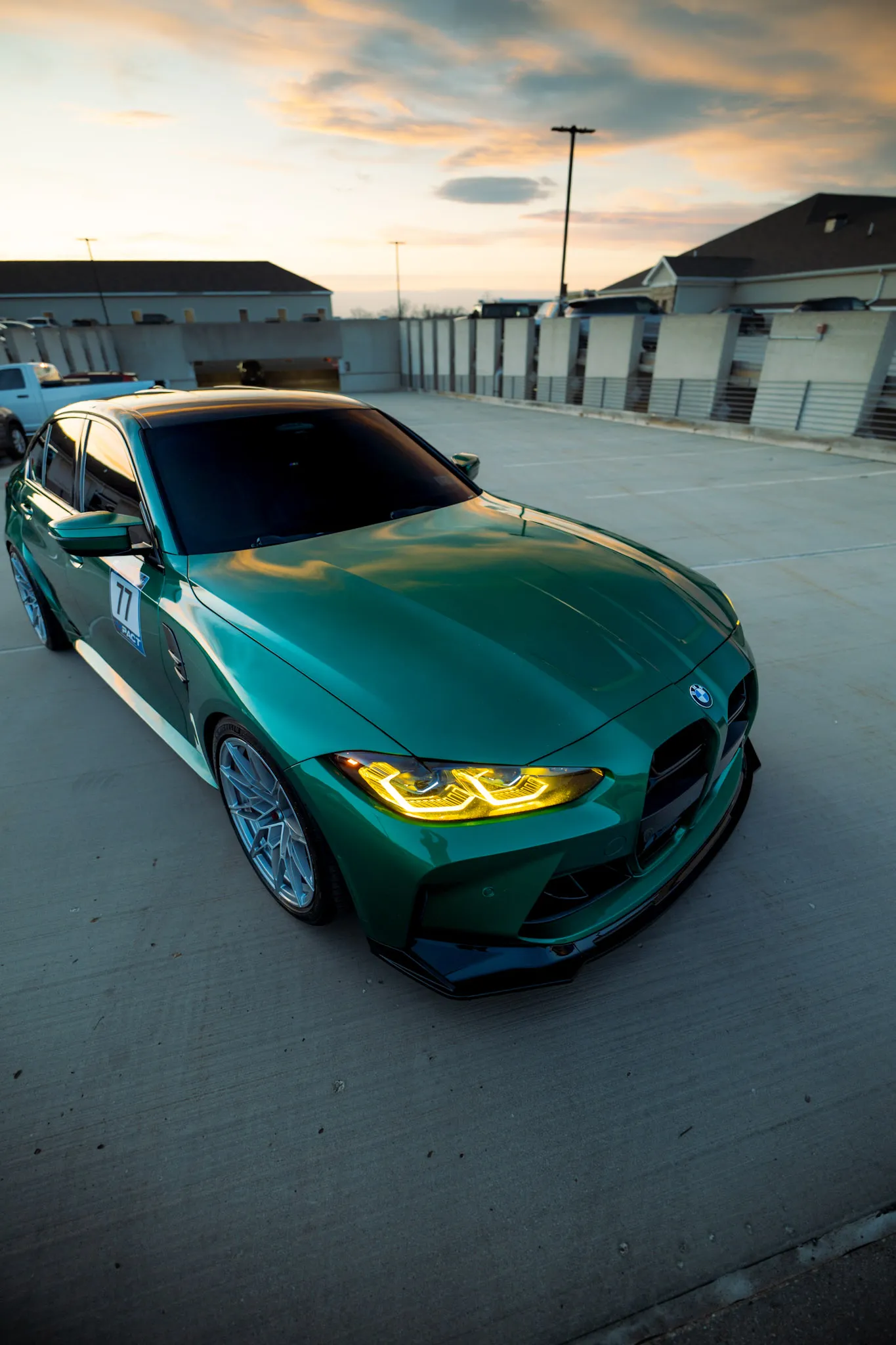 Green BMW sedan with glowing yellow headlights at sunset on a rooftop parking deck, wide-angle front view