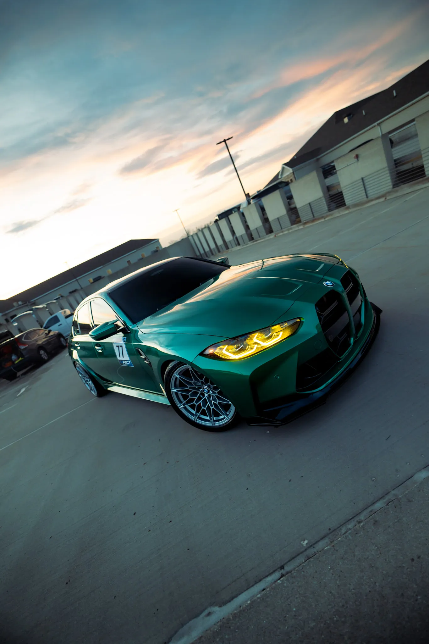 Tilted low-angle shot of a green BMW sedan with a 77 decal and lit yellow headlights under a sunset sky