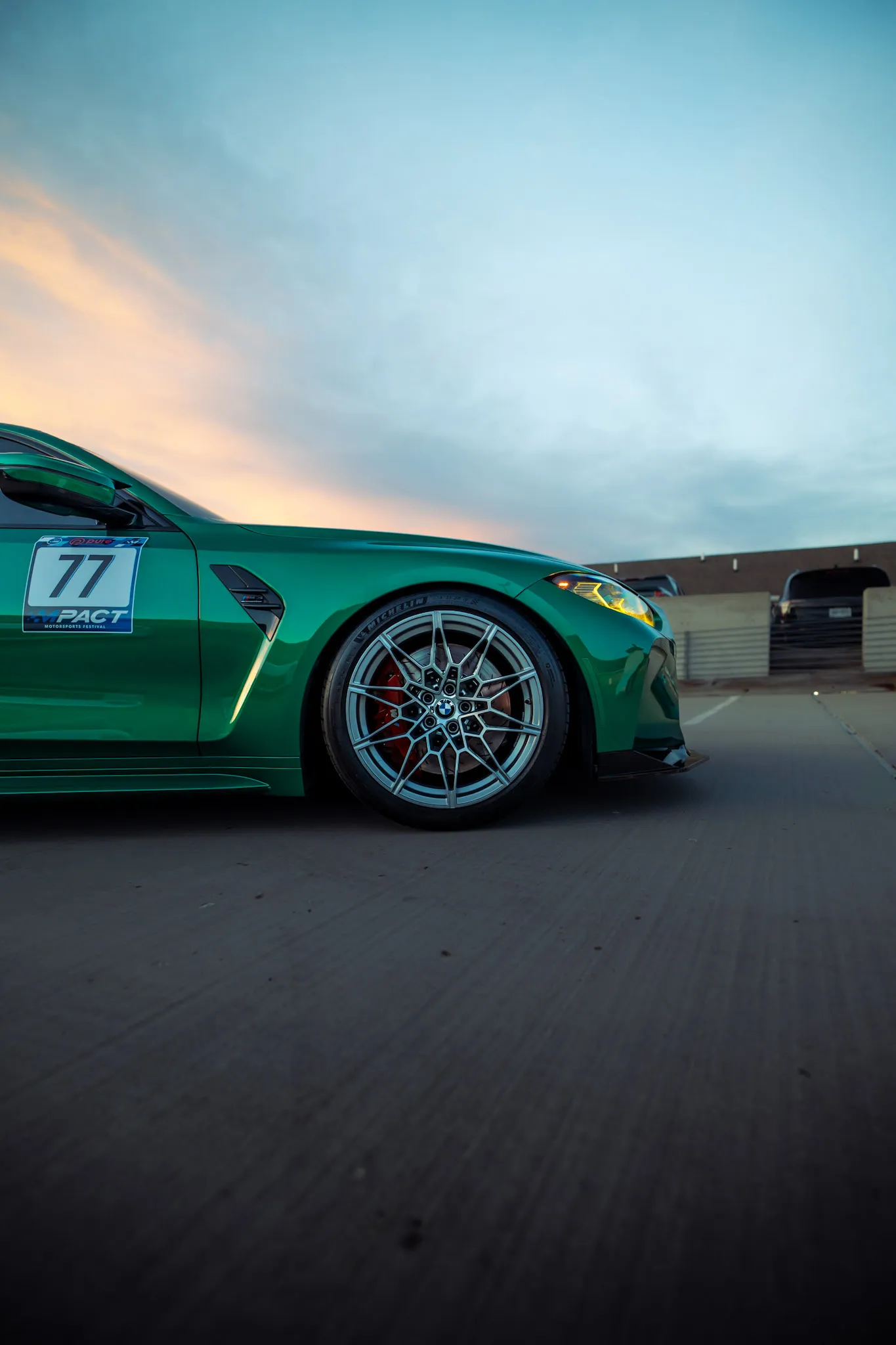 Ground-level side view of a green BMW front wheel, red caliper and 77 race number decal against a sunset sky