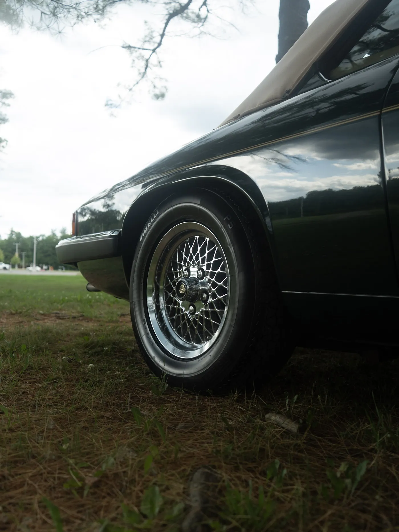 Low view of a black Jaguar's front wire wheel and Pirelli tyre, tan soft top above, under a cloudy sky