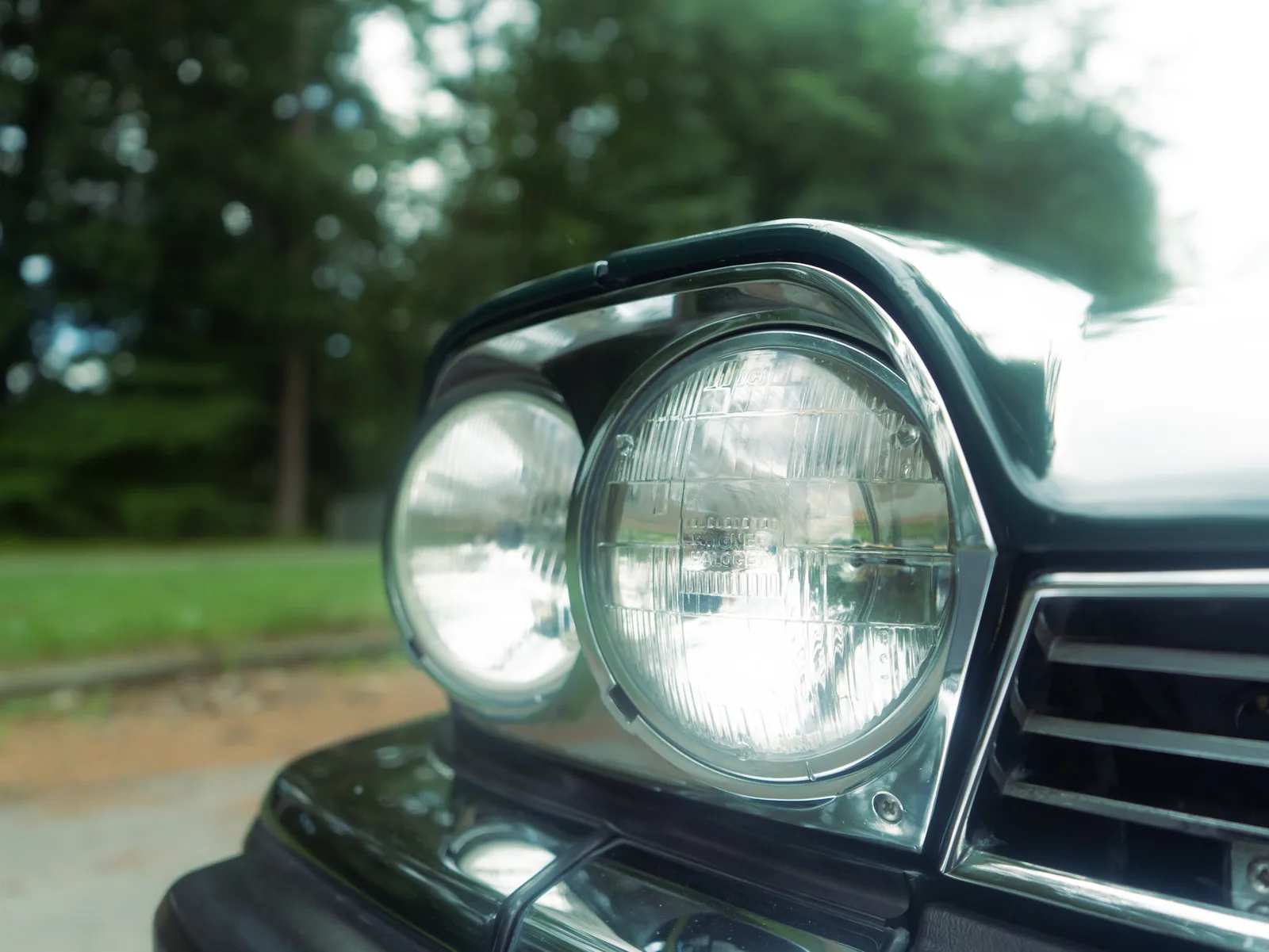 Close-up of the twin round chrome-rimmed headlights on the front of a dark Jaguar, trees blurred behind