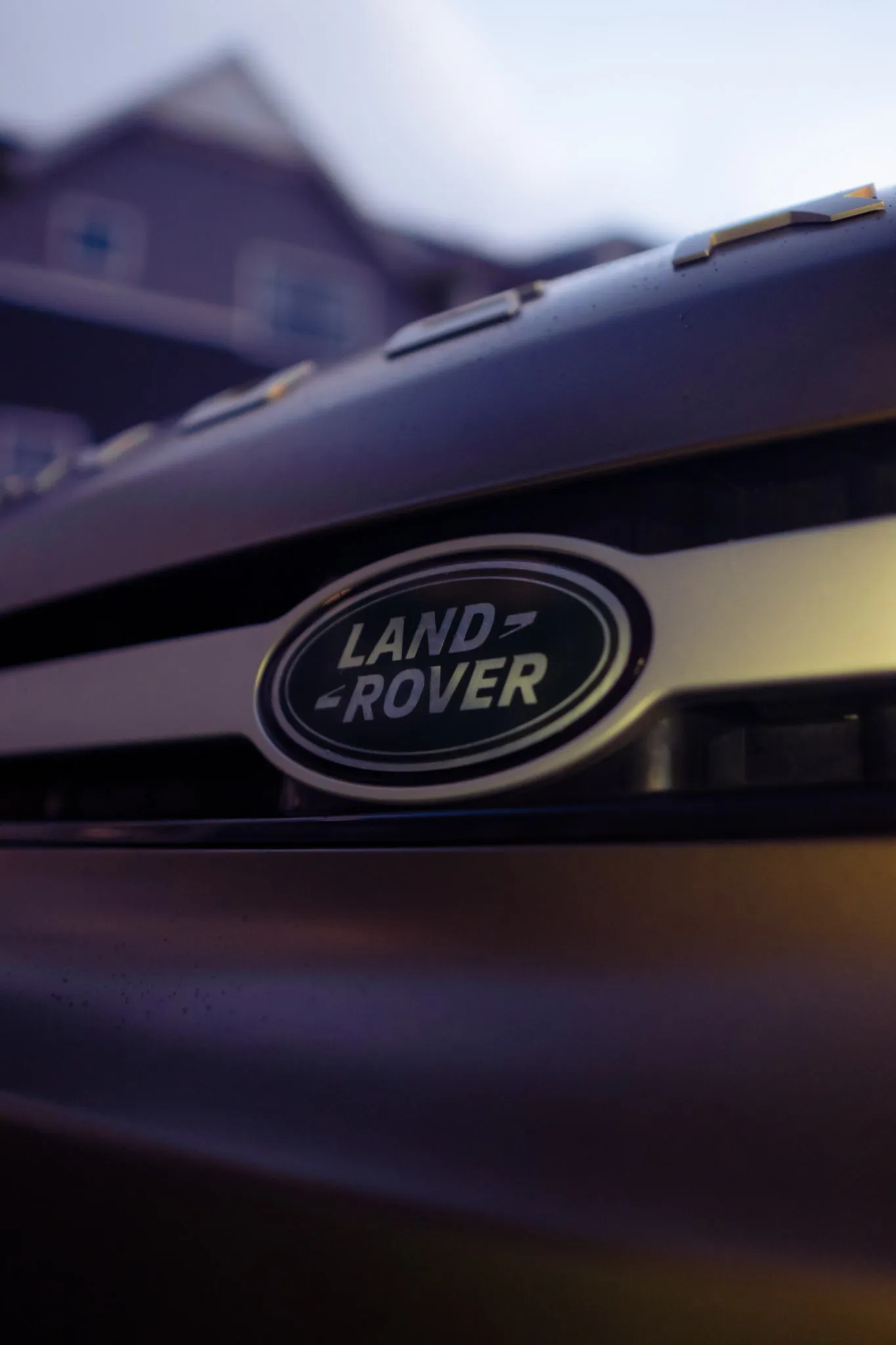 Macro close-up of the oval Land Rover badge on a Defender grille with a blurred house in the background