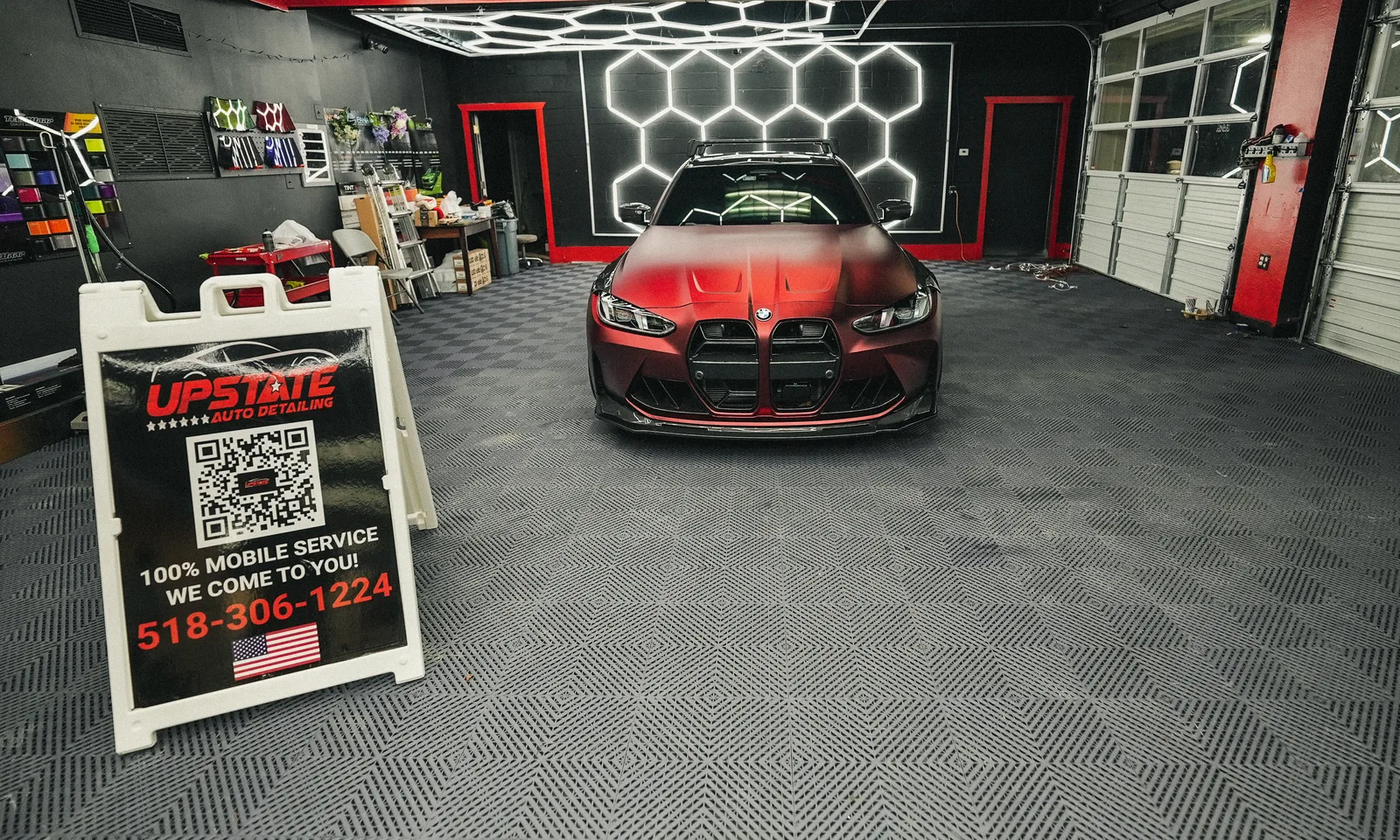 Matte red BMW parked under hexagon ceiling lights with an Upstate Auto Detailing A-frame in the foreground