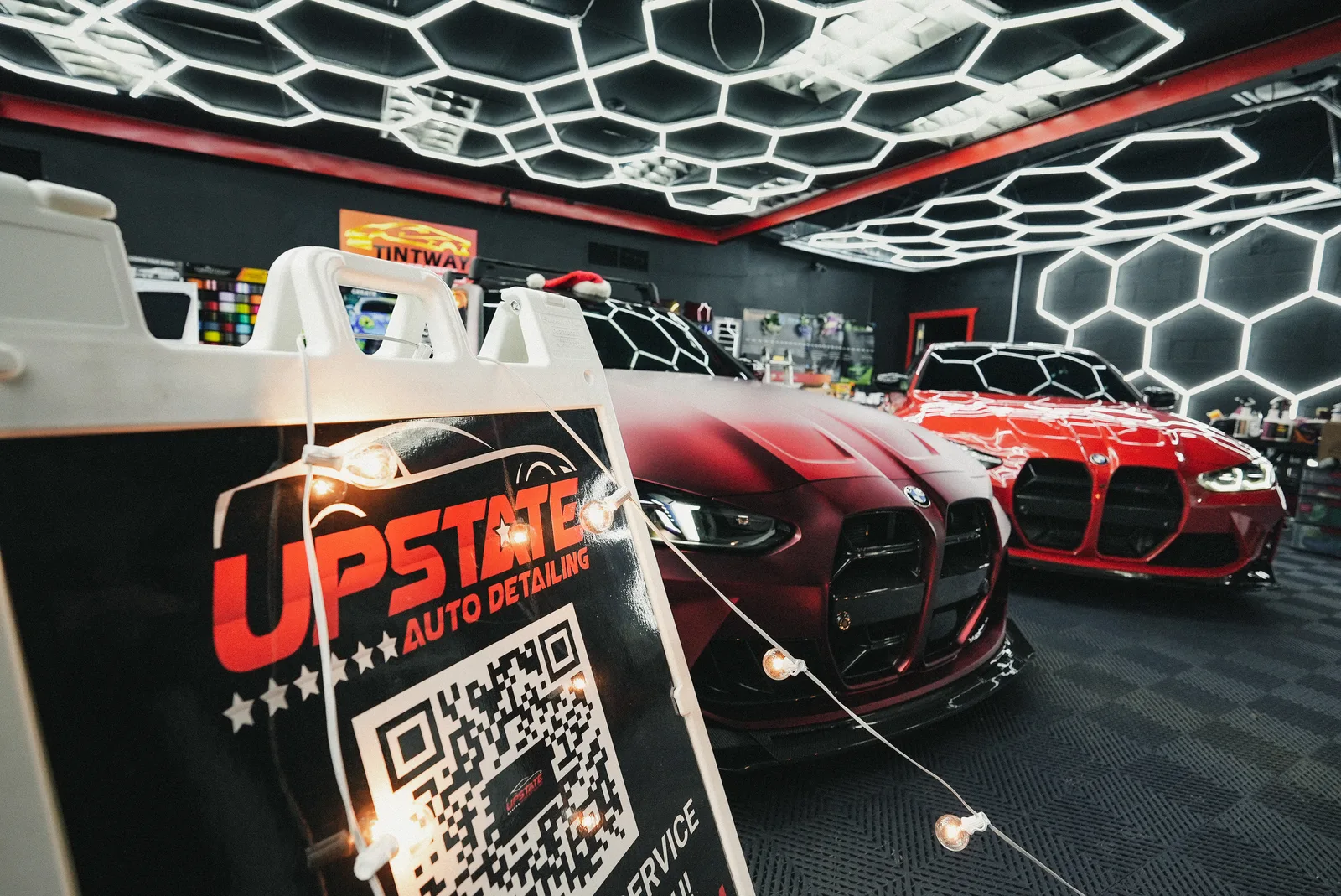 Matte red and gloss red BMWs under hexagon ceiling lights behind an Upstate Auto Detailing A-frame sign