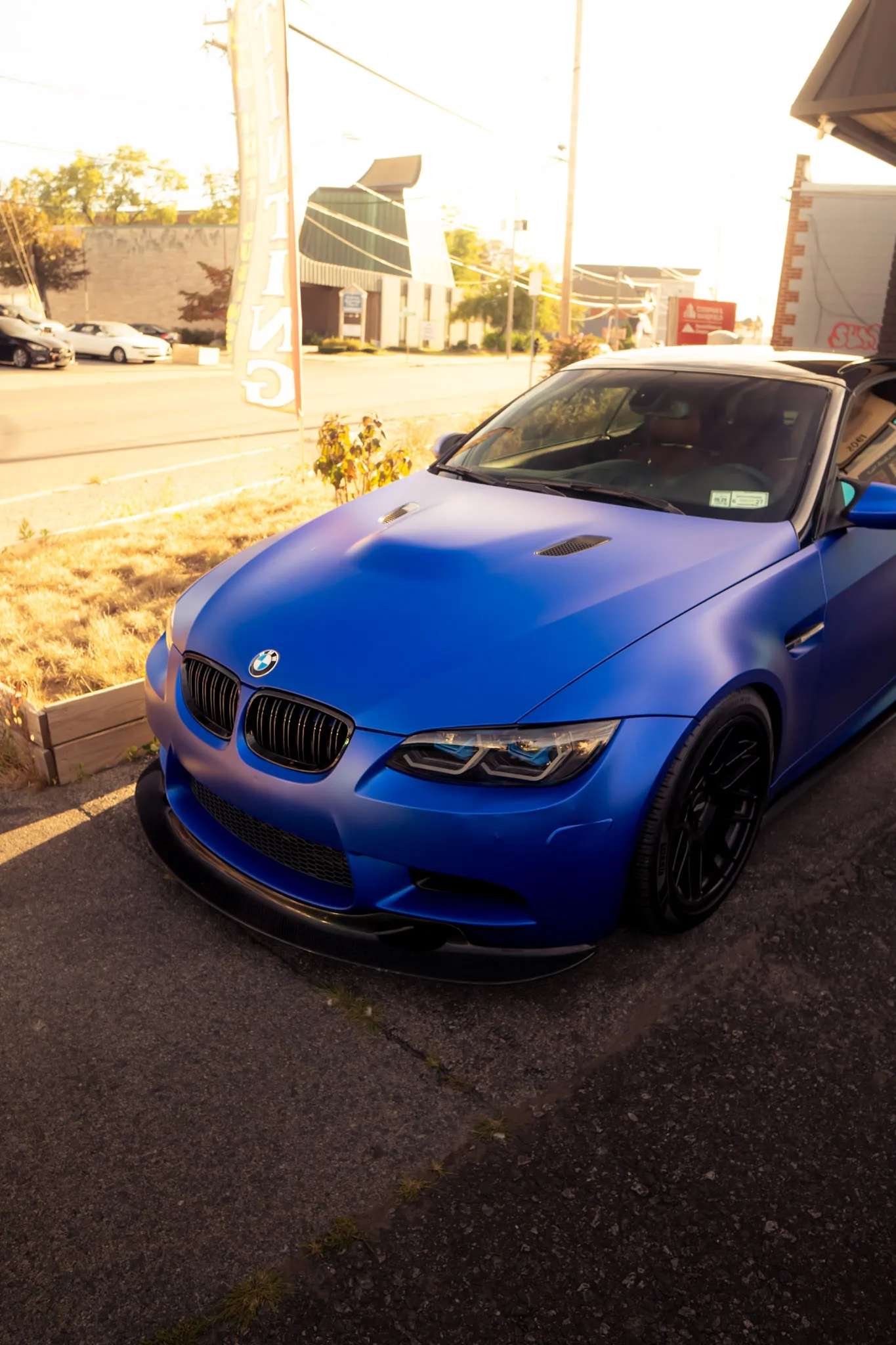 Satin blue BMW with a vented hood and black wheels parked by a street in late sun, banner flag behind
