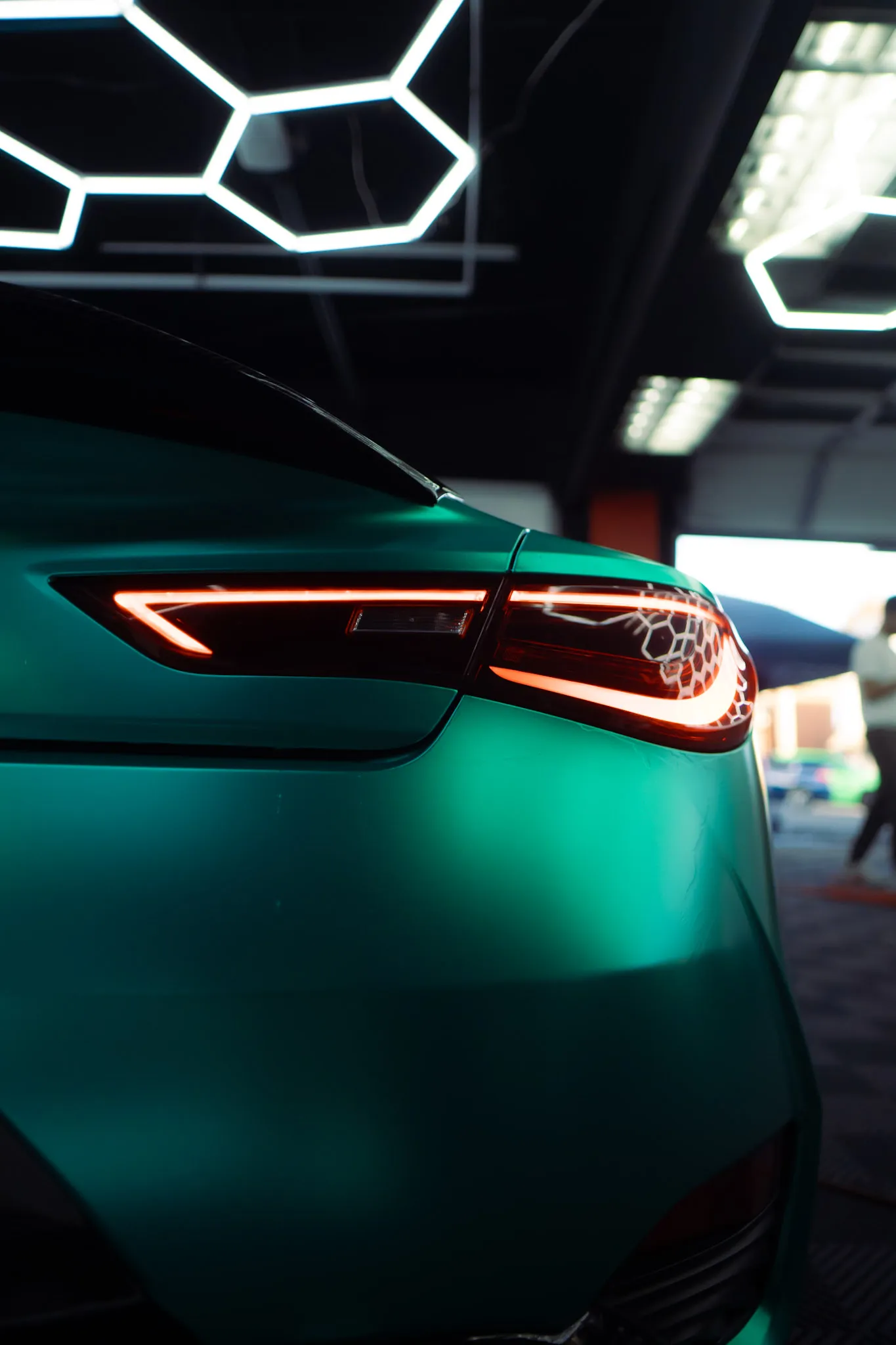 Close-up of a lit red taillight on a matte teal coupe, hexagon lights overhead and an open bay door behind