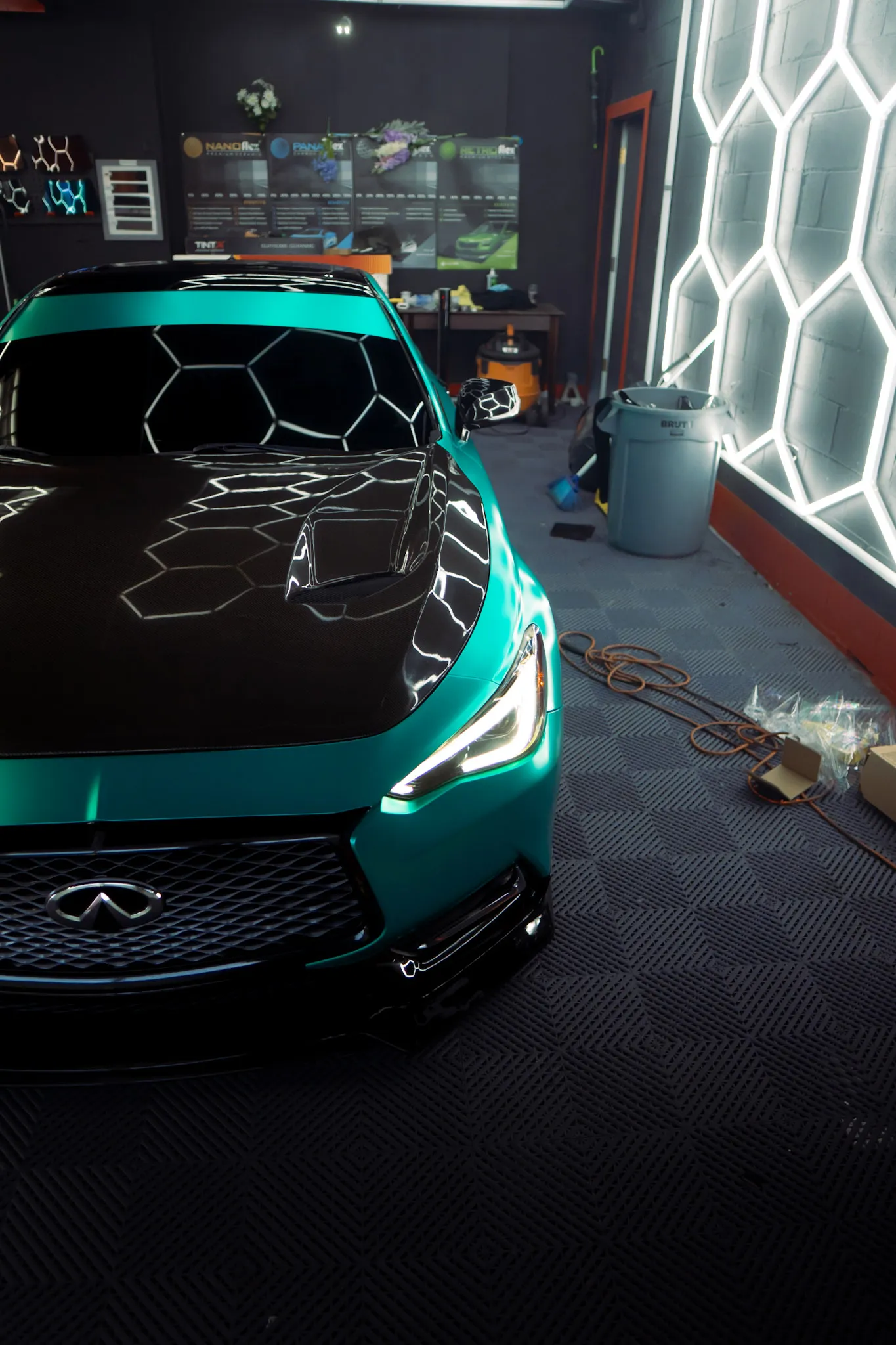 Front of a matte teal Infiniti with a glossy black hood, hexagon wall at right, trash bin and cords on the floor