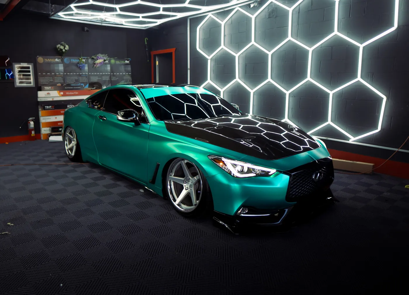 Teal Infiniti coupe with tinted windows and a black hood under hexagon lights in a tint shop