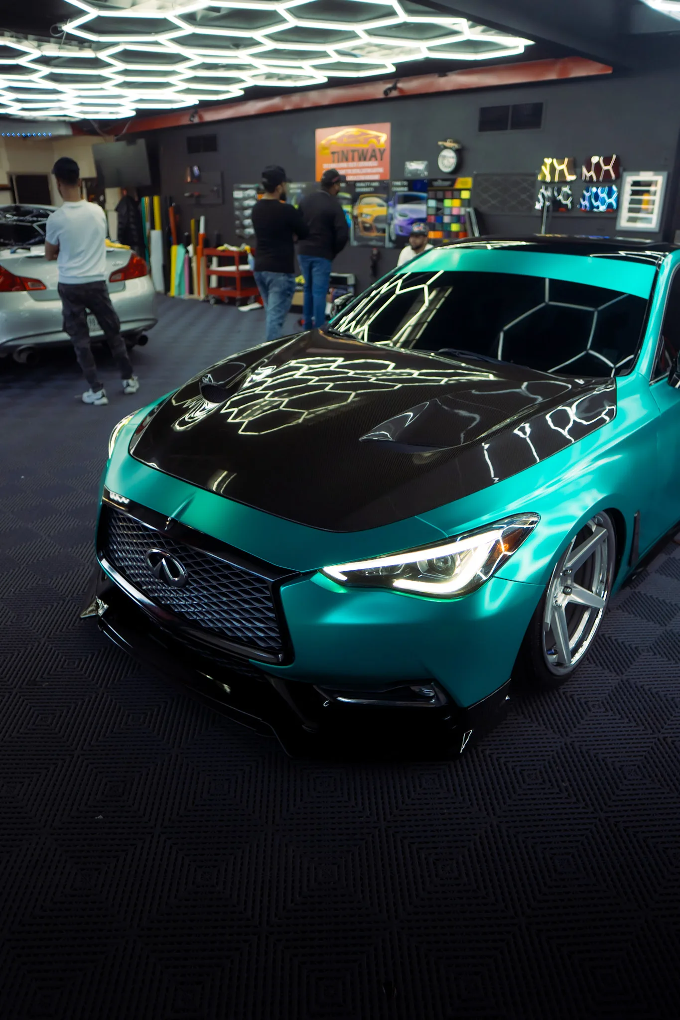 Teal Infiniti coupe with a black hood and tinted windows on the floor of a tint shop under hexagon ceiling lights