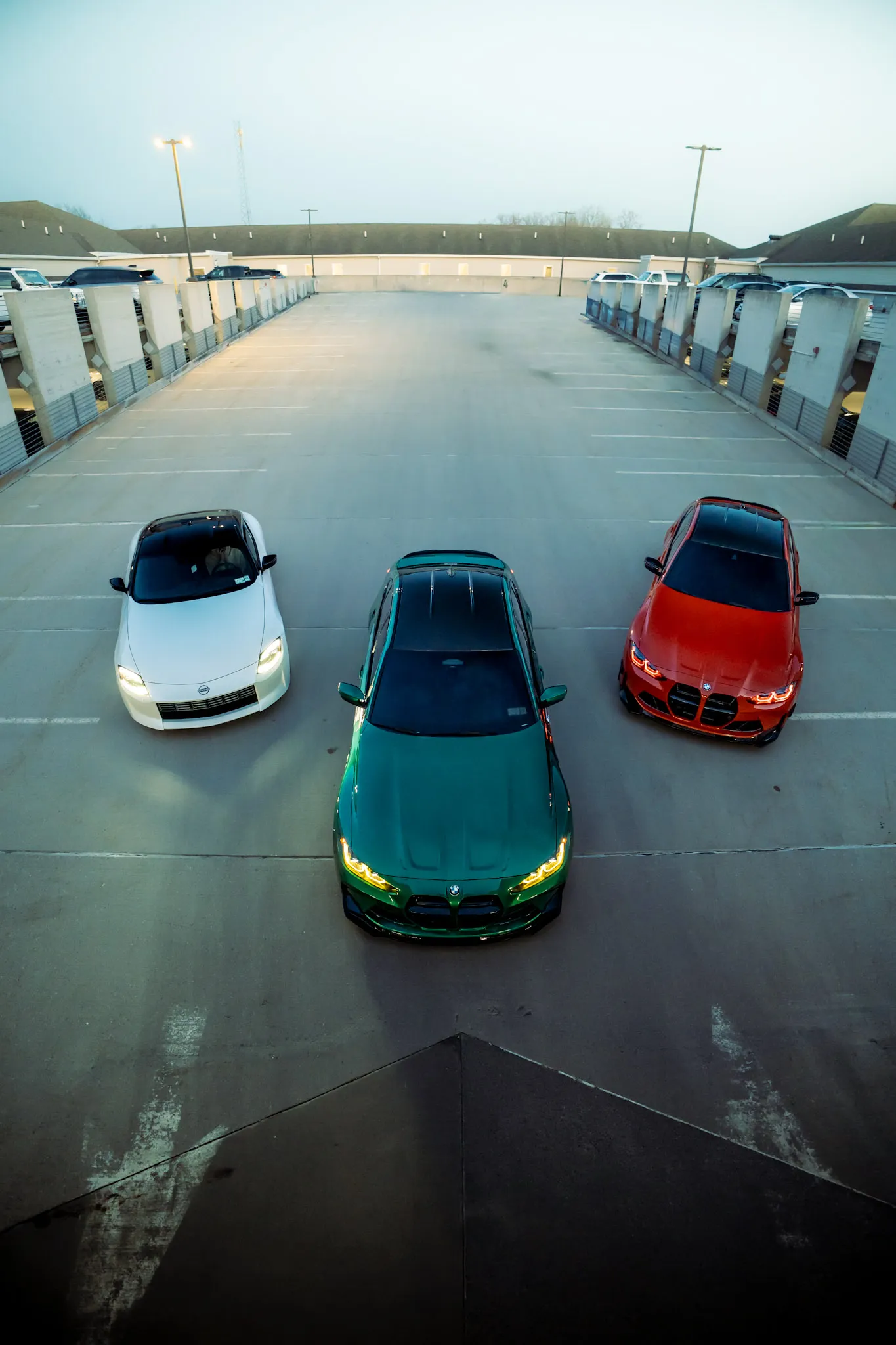 Overhead view of a white Nissan, a green BMW and an orange BMW parked side by side on a rooftop parking deck at dusk