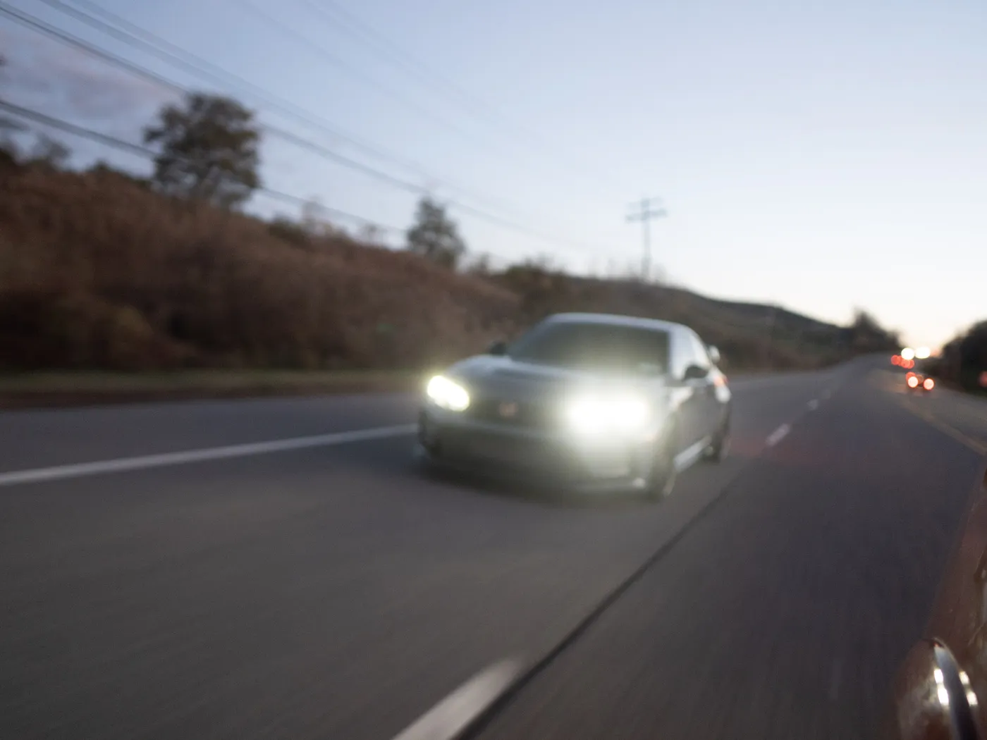 Motion-blurred gray Honda Civic Type R at dusk, headlights glowing, bare trees on the hill to the left