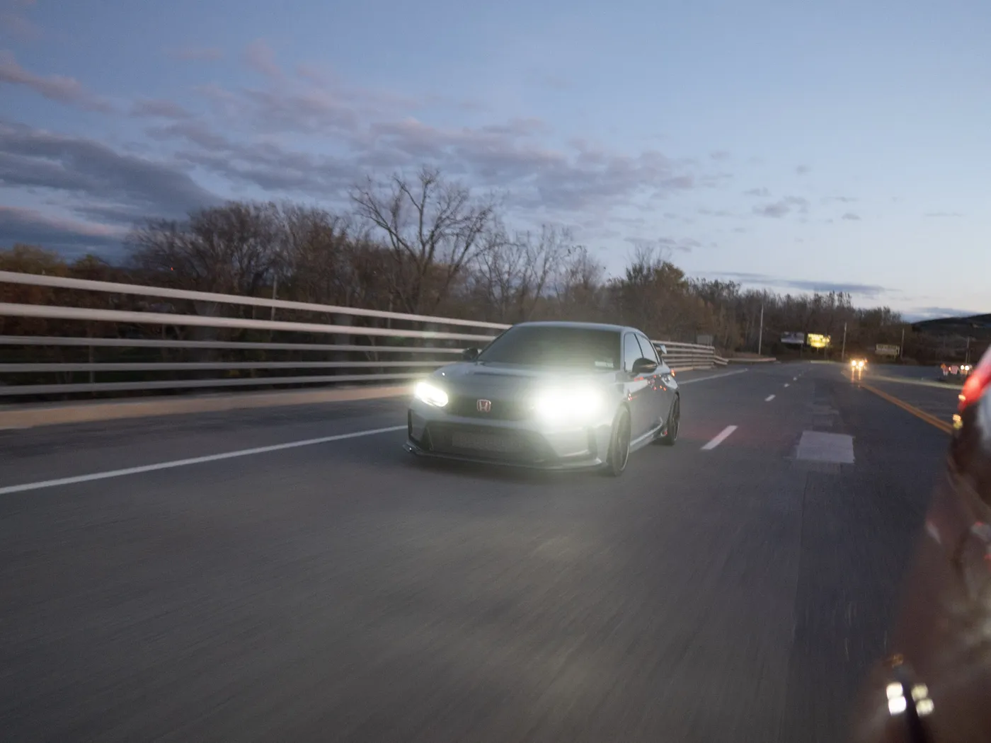Gray Honda Civic Type R crossing a bridge at dusk, guardrail and bare trees under a cloudy sky