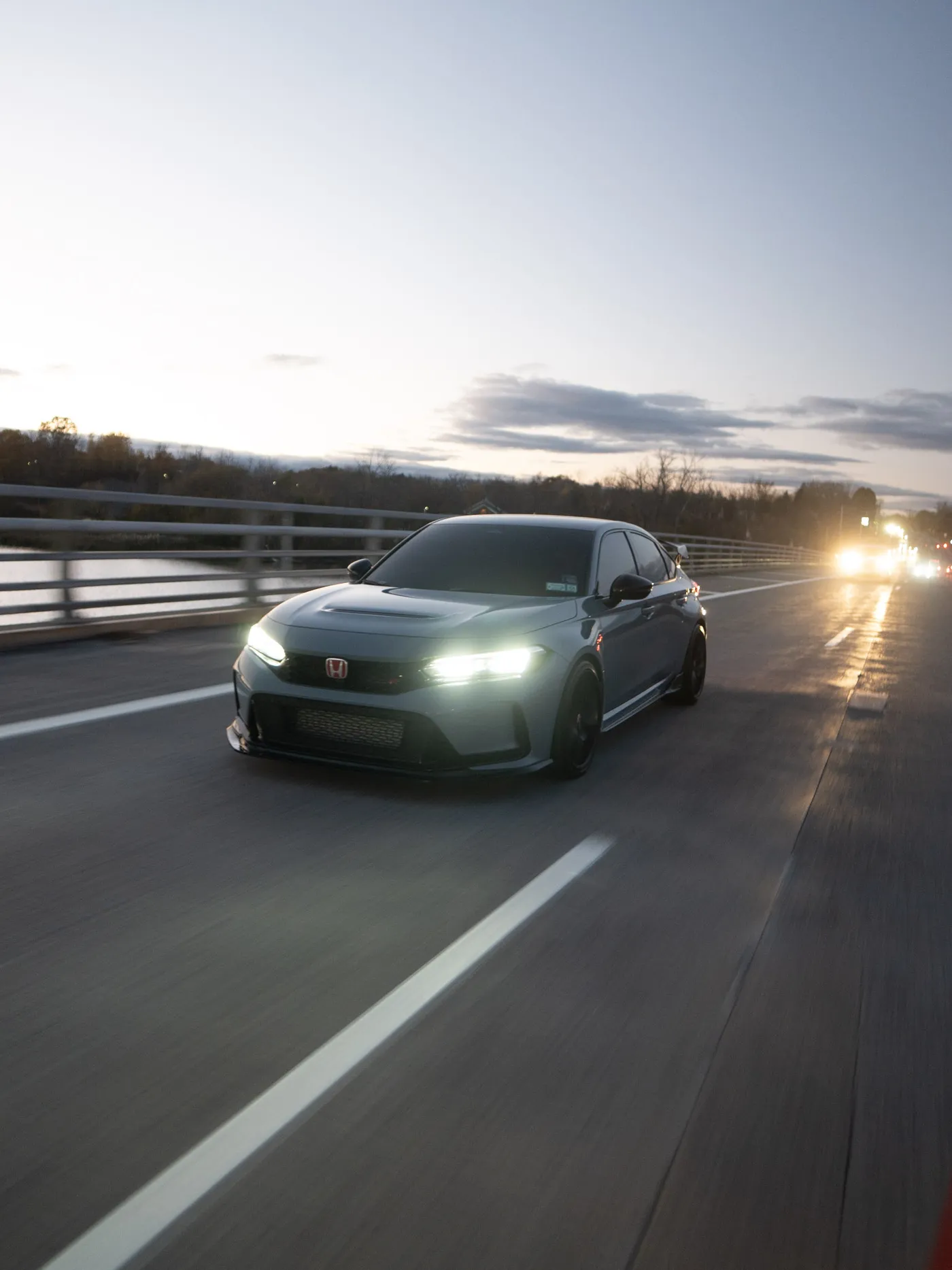Gray Honda Civic Type R on a bridge over water at sunset, headlights on and a line of traffic lights behind