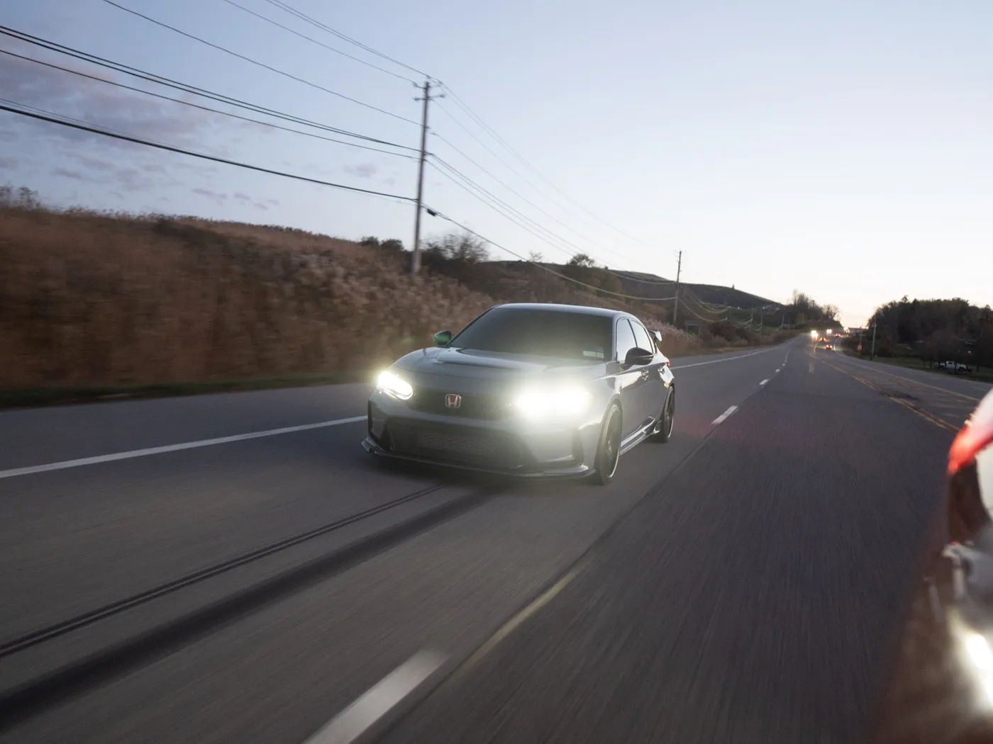 Gray Honda Civic Type R with headlights on, driving a rural highway past a dry grass hillside at dusk