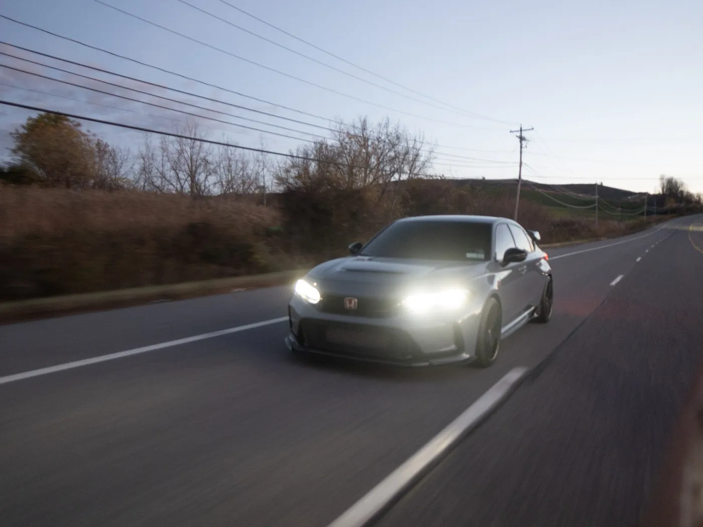 Gray Honda Civic Type R front three-quarter on an open highway at dusk, bare trees and hills behind