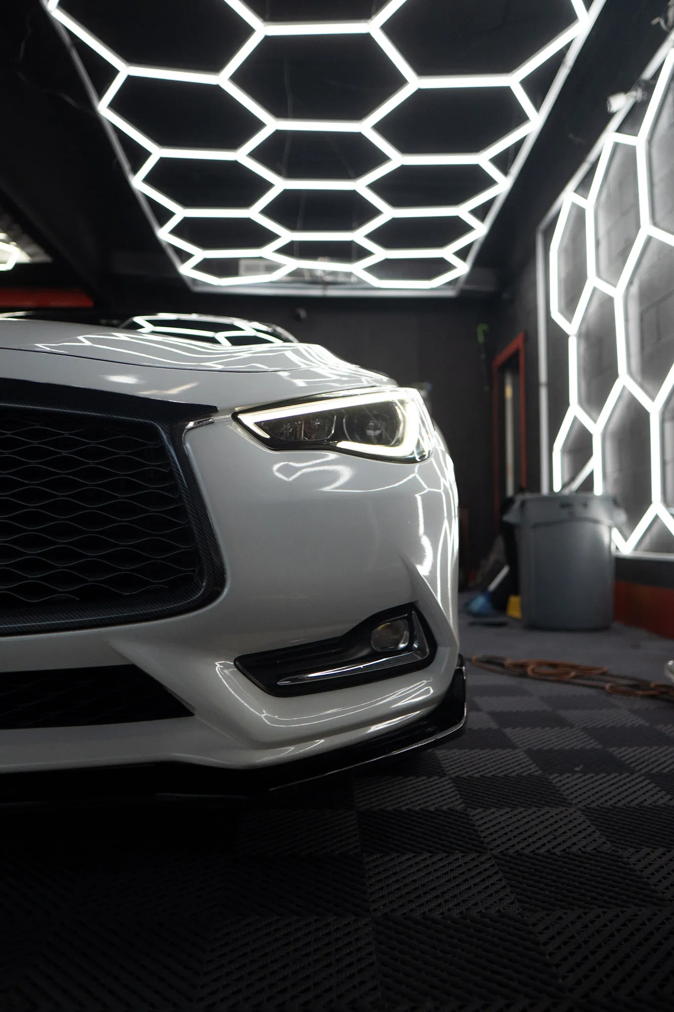 White coupe front end with headlight lit, parked under a hexagon light ceiling next to a lit hexagon wall