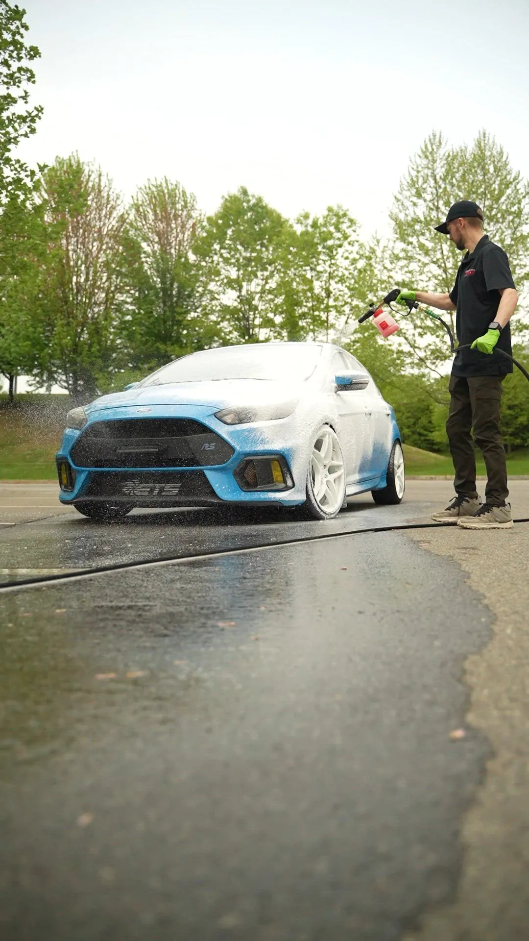 Detailer spraying snow foam from a foam cannon onto a blue Ford Focus RS, low wide shot of wet pavement