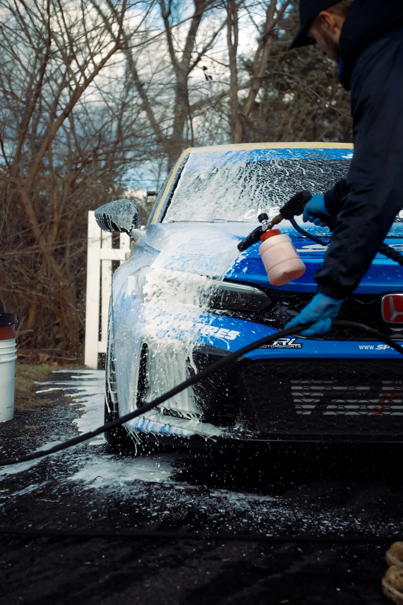 Detailer spraying thick foam from a foam cannon onto the front of a blue Honda Civic outdoors