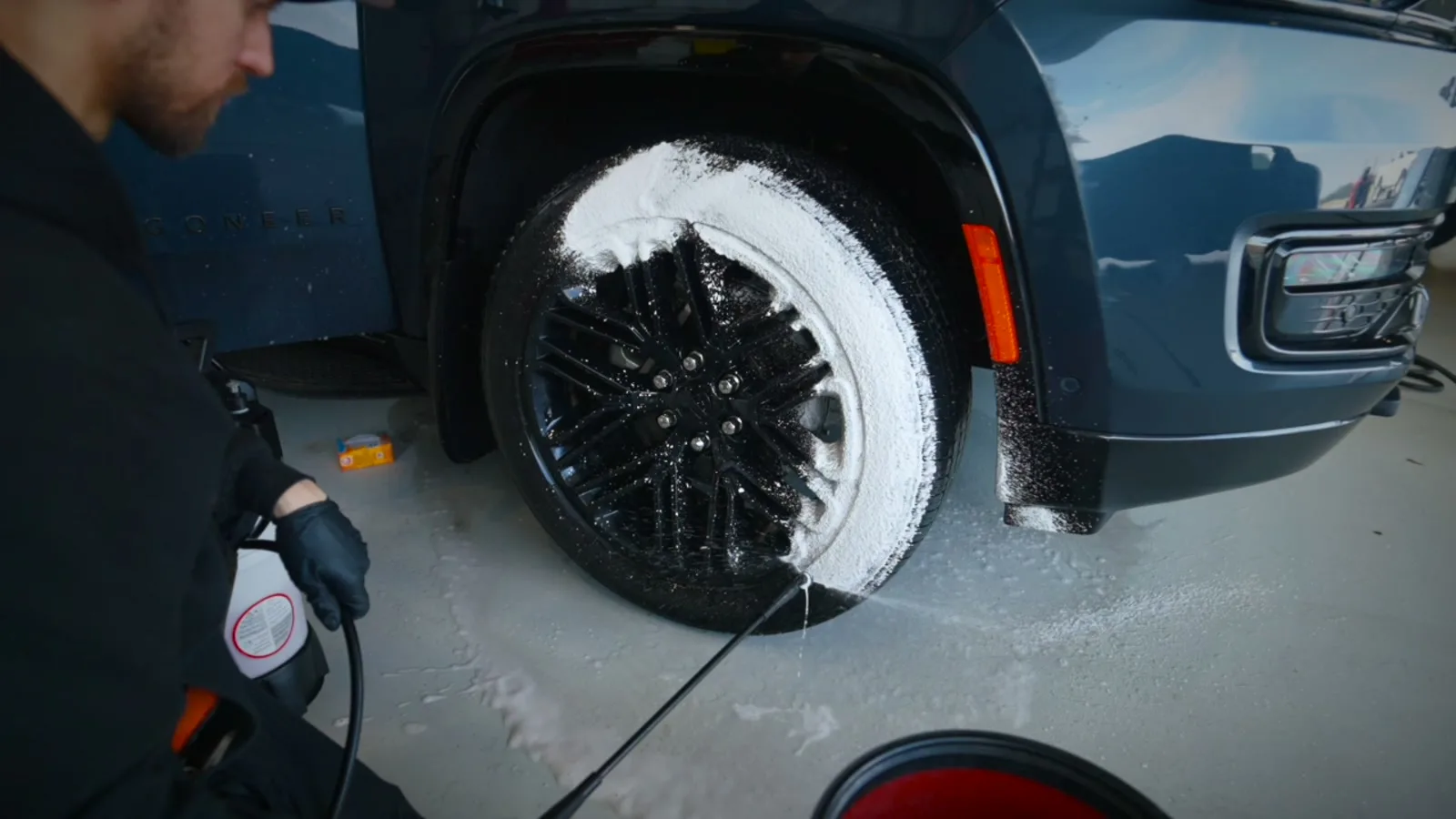 Detailer spraying foam onto the black multi-spoke wheel and tyre of a dark blue SUV