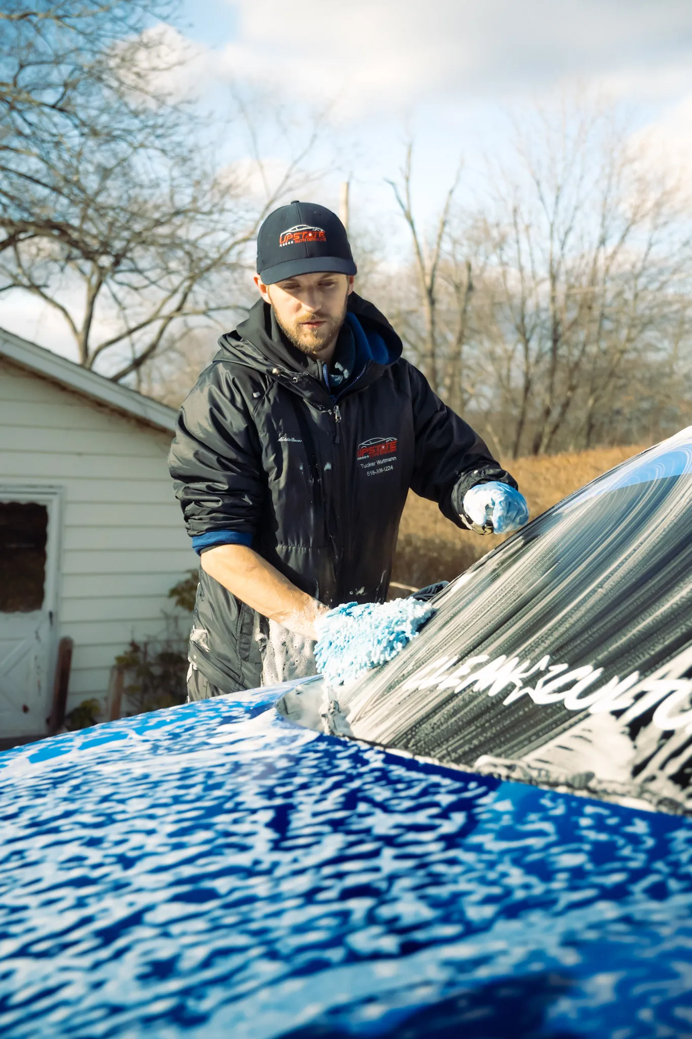 Detailer in a branded cap and jacket hand washing a foam-covered blue car windshield with a wash mitt