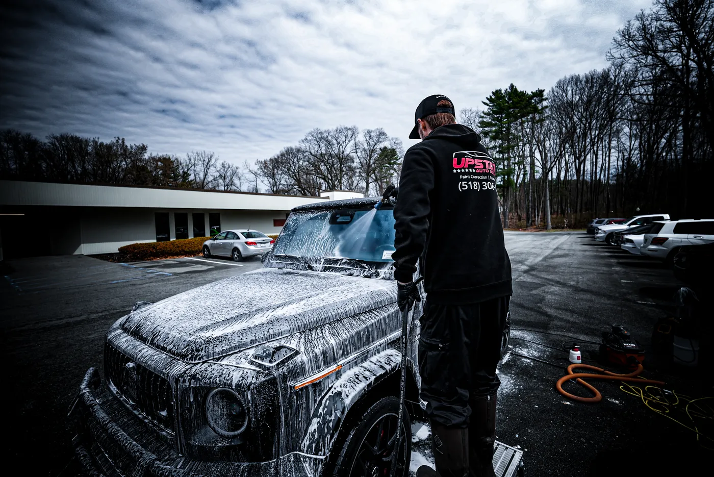 Detailer in an Upstate hoodie rinsing a foam covered dark SUV with a pressure washer wand in a parking lot