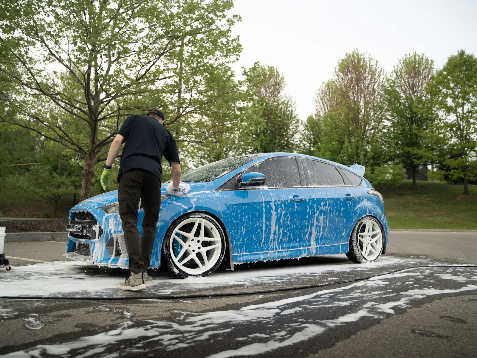 Detailer hand washing the foam-covered front bumper of a blue Ford Focus RS in an outdoor parking lot