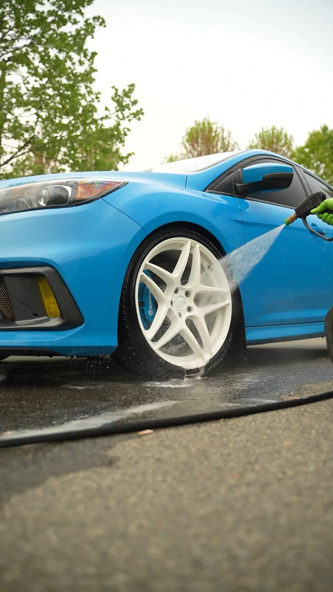 Gloved hand pressure-washing the white front wheel and blue Brembo caliper of a blue Ford Focus RS